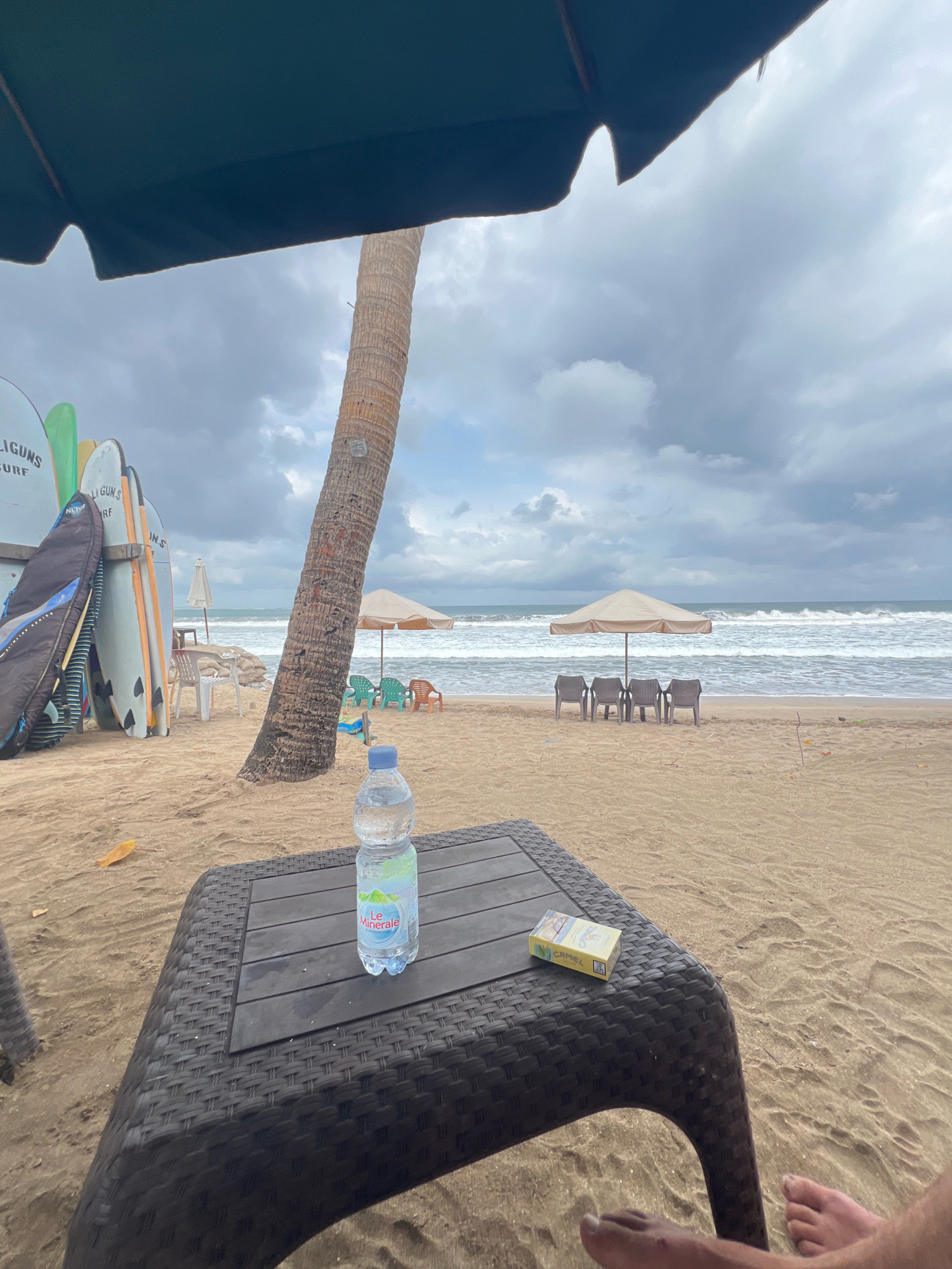 The image captures a beach scene in Kuta, Indonesia, on an overcast day. In the foreground, a dark brown, woven plastic table holds a bottle of "Le Minerale" water and a pack of Camel cigarettes. To the right, two bare feet are visible, suggesting someone is relaxing. The table is positioned on a sandy beach. In the midground, a palm tree trunk stands prominently, and behind it, a row of surfboards are propped against a low wall. Beyond the surfboards, several beach chairs and umbrellas are set up on the sand, with the ocean in the background, displaying moderate waves. The sky is filled with heavy, gray clouds.