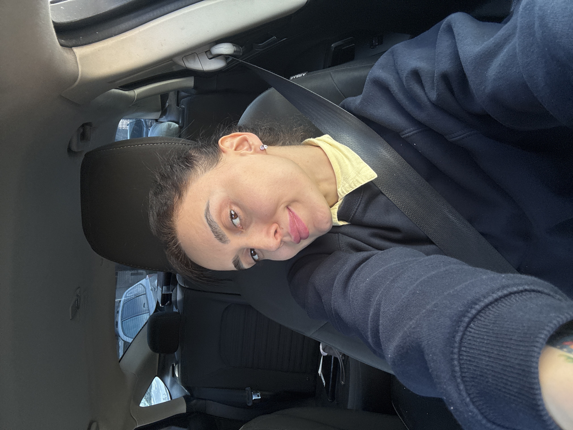 A selfie shows a woman wearing a yellow shirt and a navy blue sweatshirt with a seatbelt across her chest, positioned in the driver's seat of a car. The car's interior is visible. Outside the car window, a vehicle is visible. The location is most likely in Tbilisi, Georgia, based on the vehicles observed outside.