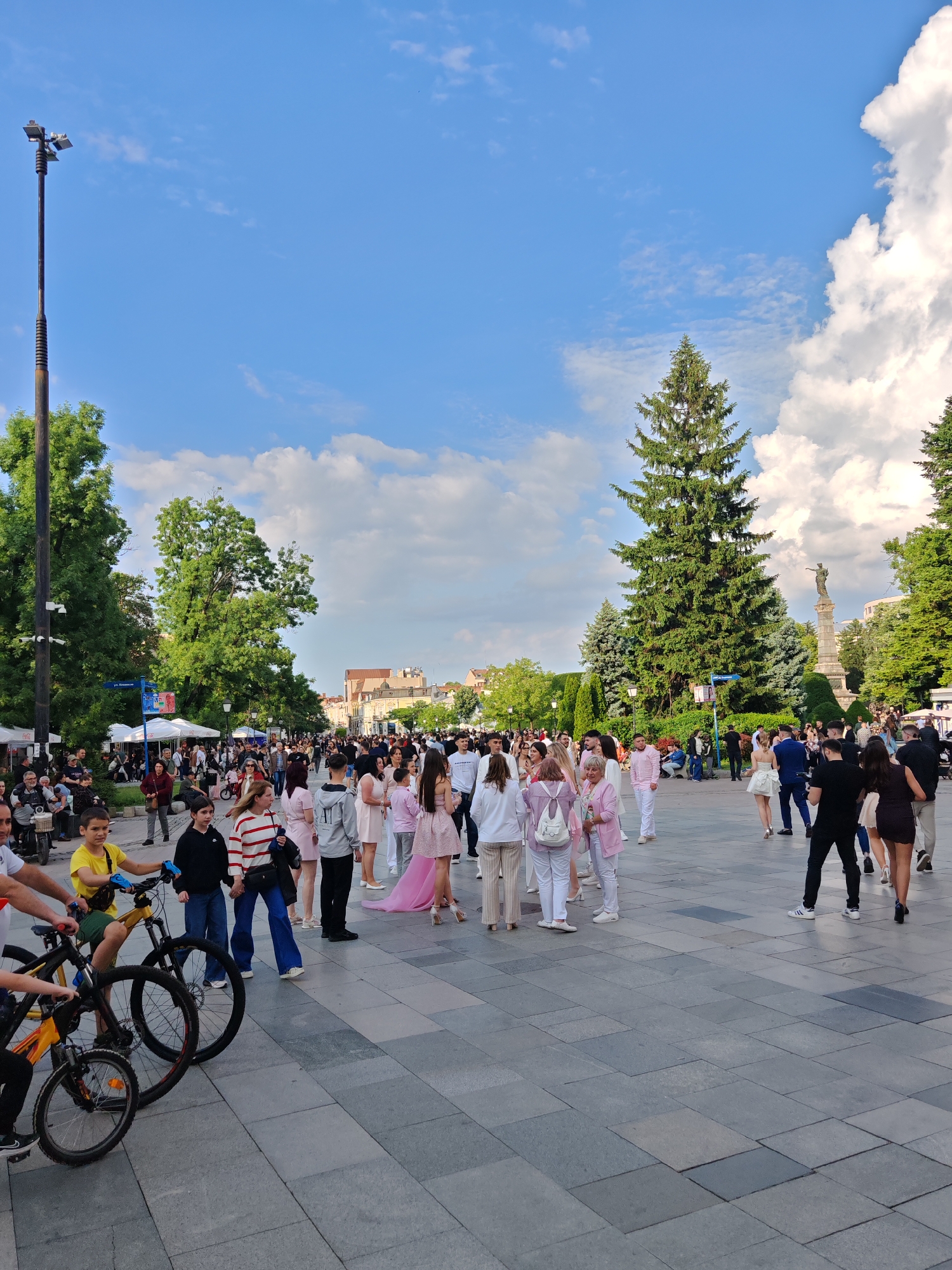 The image shows a public square in Ruse, Bulgaria, filled with people. A large group, mostly dressed in white and pink, is gathered in the center of the frame. Some individuals are also wearing casual clothing. Bicycles are visible in the lower left corner. The ground is paved with grey stone tiles. In the background are trees and a monument, along with buildings. A tall light pole stands on the left edge of the image. The sky is blue with scattered white clouds. The overall scene depicts a gathering or event taking place in the city center.