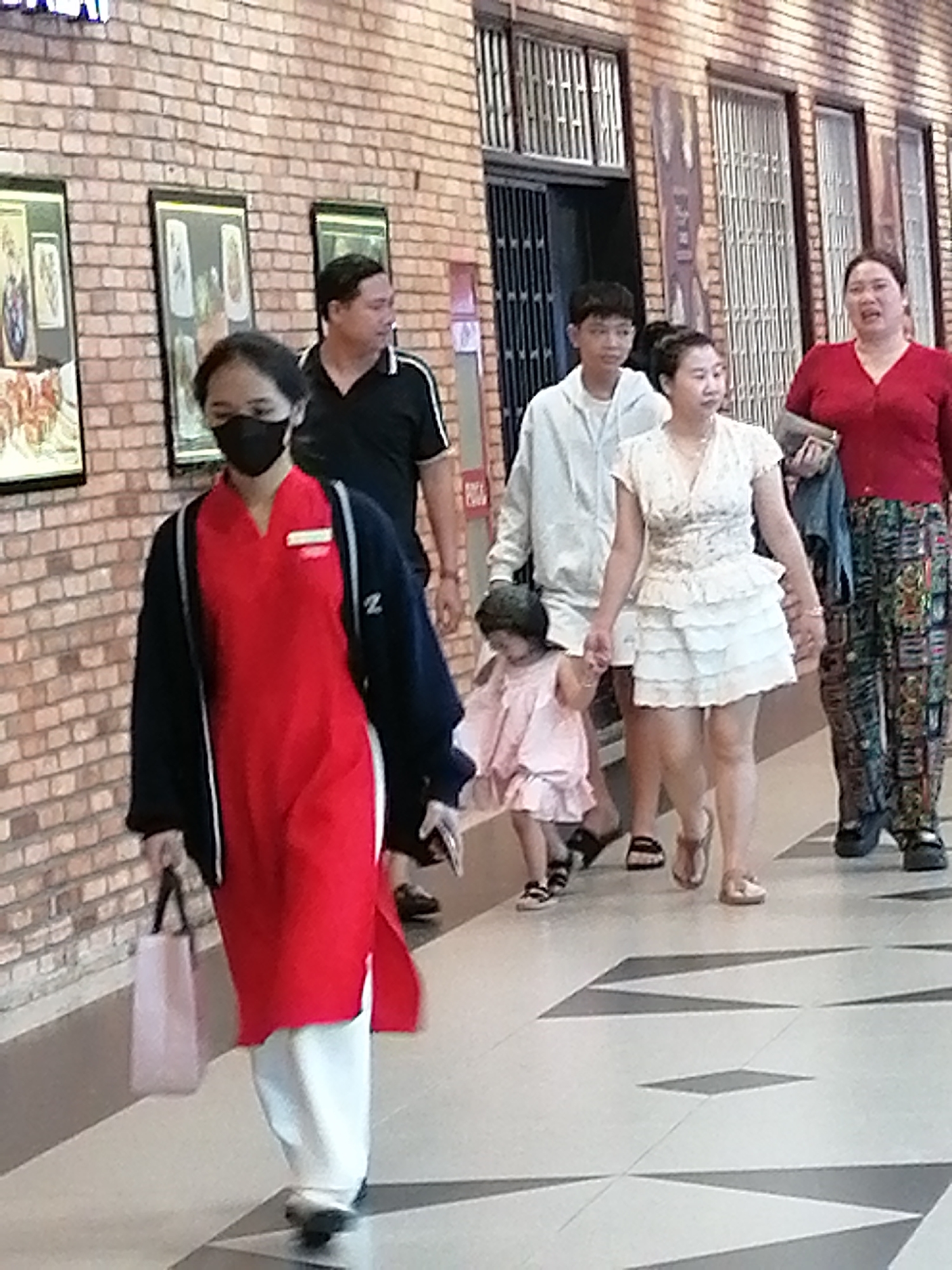 The image depicts a group of six individuals walking along a tiled pathway next to a brick wall. The individuals include two adult women, two adult men, and two young children. The woman in the foreground is wearing a red tunic and white pants, with a black jacket over her shoulders and a black mask covering her face. She is carrying a light pink shopping bag in her left hand. To her right, a woman in a white, ruffled dress is holding the hand of a young child in a pink dress. Behind them, a young man in a light grey hoodie walks, and further back, an adult man in a black polo shirt walks with another adult woman wearing a red top and patterned pants. The environment appears to be an indoor walkway, possibly within a market or shopping area, given the presence of shops with metal grilles visible on the wall in the background. The lighting suggests an indoor setting, likely during daylight hours. The overall scene captures a moment of people moving through a public space.