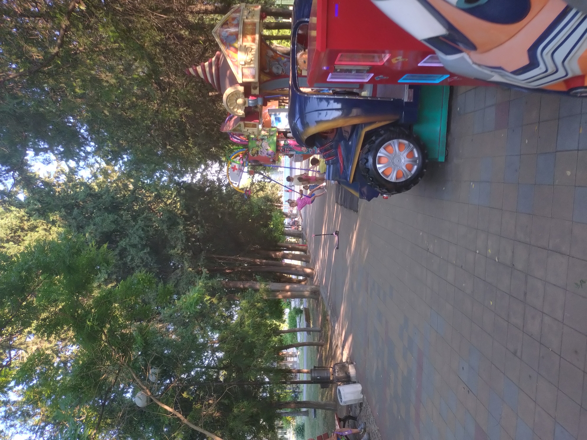 The image shows a row of amusement rides and attractions along a tree-lined pathway. The rides appear to be designed for children, with colorful facades and cartoonish characters. One ride, in the foreground, is shaped like a blue and red vehicle with a large orange-rimmed wheel. Several children are visible playing on or near the rides.
The scene is set outdoors in what appears to be a park or fairground. Tall trees with dense green foliage line the pathway, casting dappled shadows on the ground. The pathway itself is paved with square tiles in a mosaic pattern. The time of day seems to be late afternoon or early evening, judging by the warm, soft light and the long shadows. The weather appears to be pleasant and sunny.
In the background, more trees and some distant structures can be seen. The overall atmosphere is one of fun and recreation. There is no visible text in the image. The location is Stara Zagora, Bulgaria.