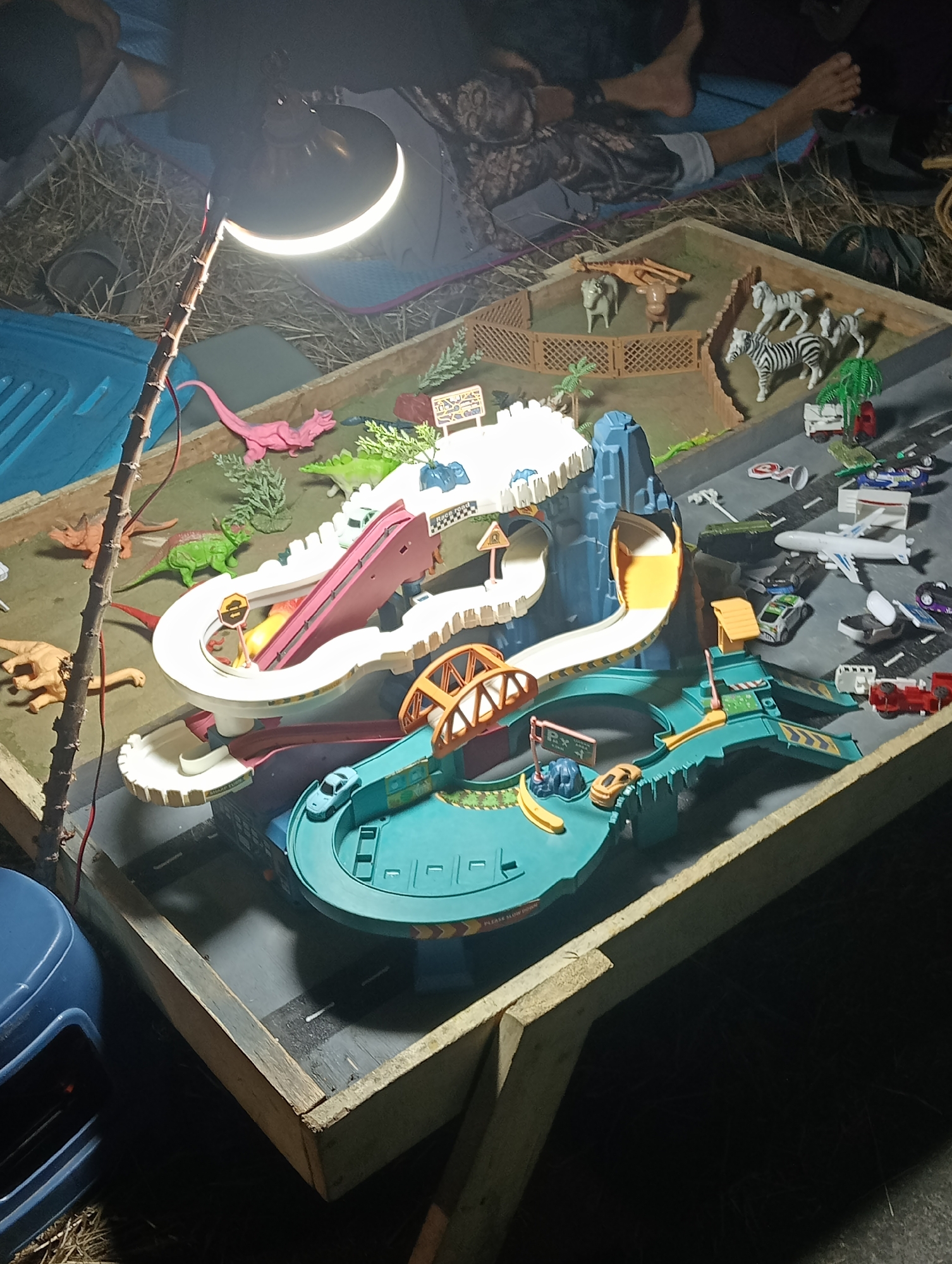 In Ngemplak Wetan, Indonesia, a diorama depicting a race track and a zoo is set up outdoors, possibly for play or display. The scene is illuminated by a bright lamp, casting shadows. In the background, two individuals, a male and a female, are partially visible, lying down on a grassy area. The diorama features various toy animals, including dinosaurs and zebras, as well as toy cars and a toy airplane. A ramp and track system for toy cars is the central element, with a large blue plastic container to the left and dried straw scattered around the setup. The diorama is enclosed in a wooden frame.
