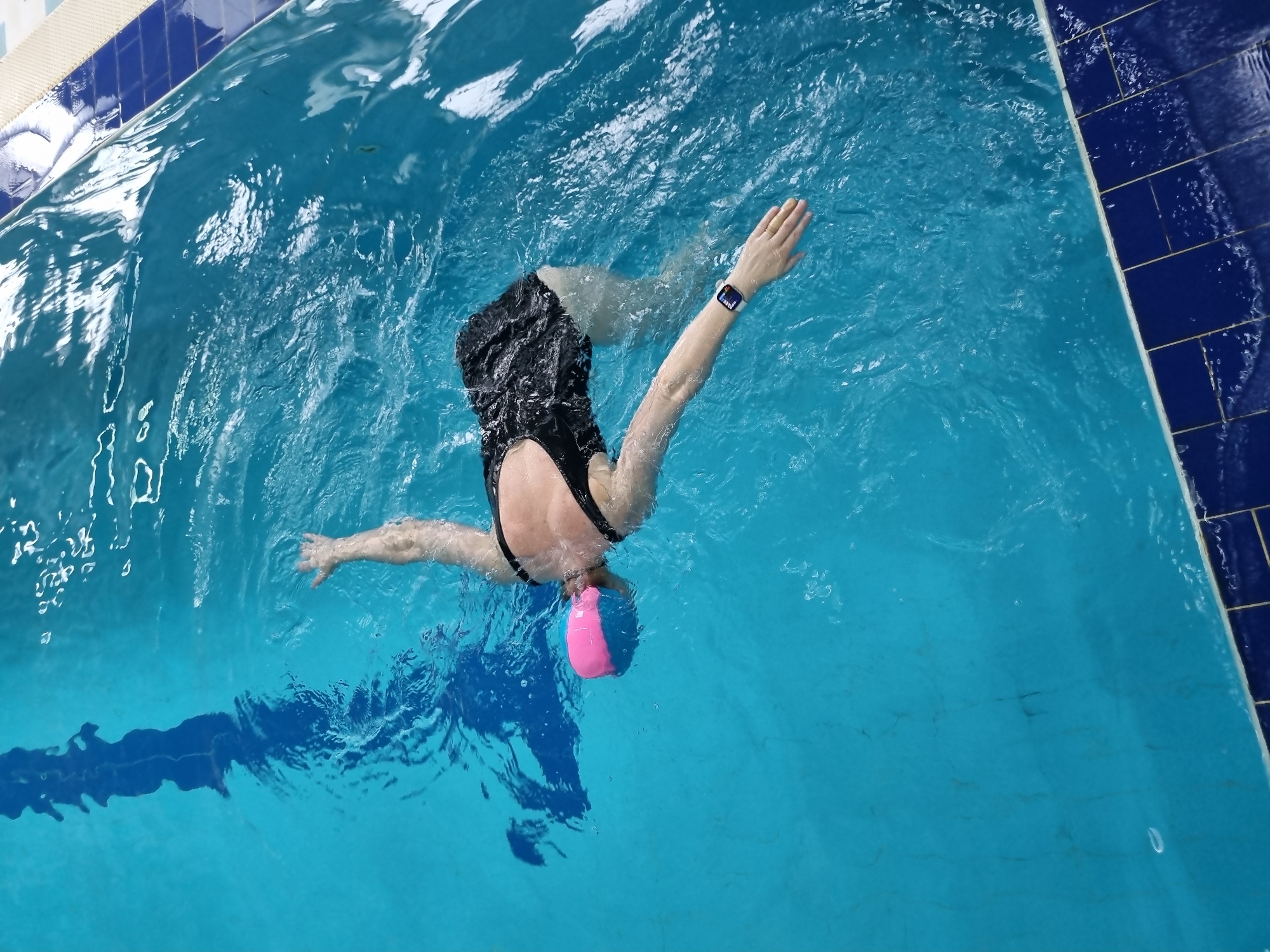A person is swimming in a pool. The person is wearing a black swimsuit and a pink and blue swim cap. The person's arms are outstretched and they appear to be swimming the breaststroke. The pool is blue and there is a lane marker in the water. The location is not specified.