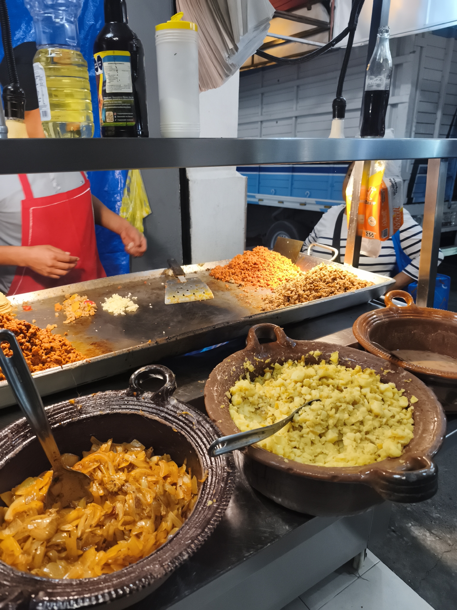 The image shows a street food stall in Mexico City, Mexico. A person wearing a red apron is preparing food on a flat-top grill, while another person in a striped shirt is seated behind the food. There are three earthenware bowls containing various fillings. One bowl contains what appears to be stewed onions, another contains diced potatoes, and the third contains a light-colored liquid, possibly a broth or sauce. A variety of condiments and liquids sit on the shelf above the food preparation area, including bottles of oil, soy sauce, and Coca-Cola. Packages of what appear to be tortillas are seen behind the grill and in front of the person in the striped shirt. The street food stall is attached to a light blue truck.