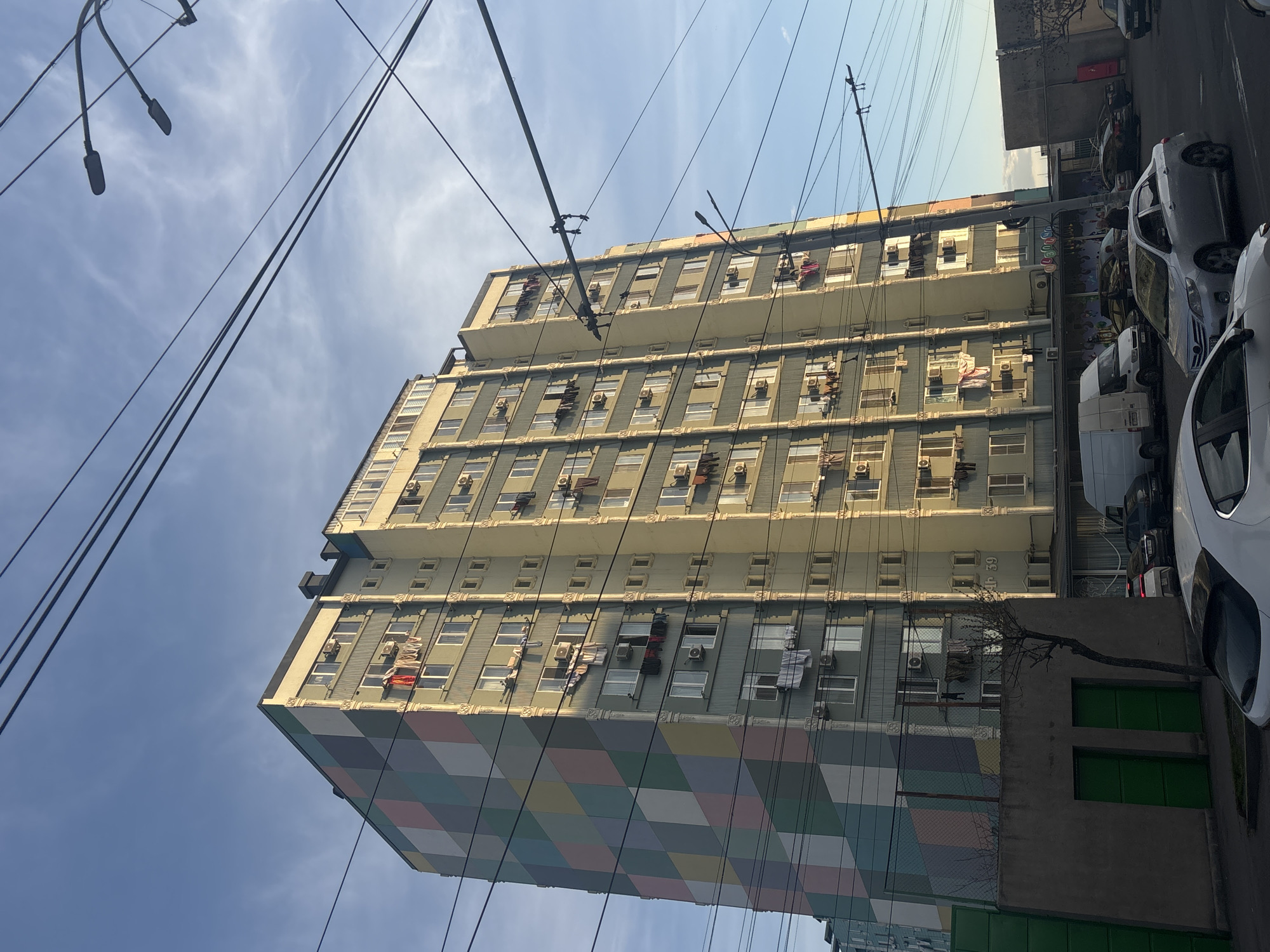 The image shows a tall apartment building in Yerevan, Armenia. The lower part of the building features a checkered pattern in pastel colors. The upper stories are painted a light gray, with numerous windows and air conditioning units. Laundry is hung out to dry on some of the windowsills. Electrical wires crisscross the sky above the building. Parked cars line the street next to the building, and a concrete structure with green doors is visible at the base of the building. A small tree branch extends across the lower right of the frame. The sky is blue with scattered clouds. The building’s facade displays the number 39.