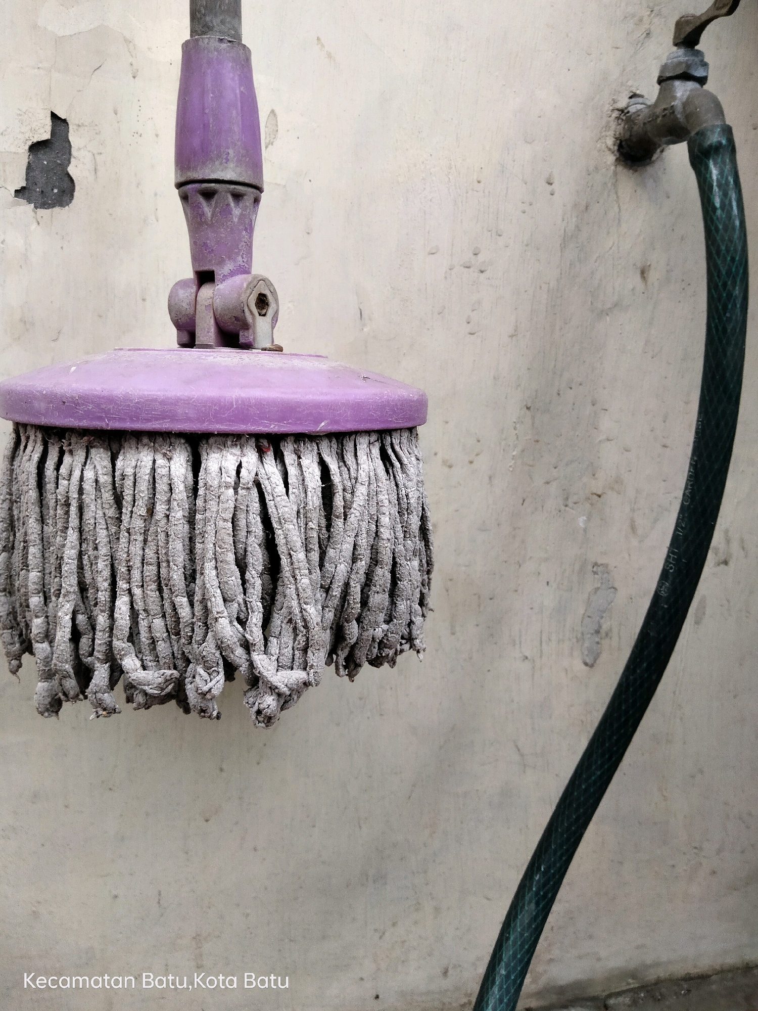 A close-up, eye-level outdoor shot depicts a worn purple mop head hanging against a textured, off-white stucco wall. The mop's plastic handle and head show signs of age and use, with some peeling paint on the upper part of the handle. Attached to the wall to the right is a tarnished metal faucet with a green, ribbed hose connected to it, curving downwards and out of view. The wall itself has patches of discolored and peeling stucco, revealing darker concrete beneath. The bottom left corner of the image contains white text that reads "Kecamatan Batu, Kota Batu," indicating the location as Batu District, Batu City, Indonesia. The lighting is even, suggesting an overcast day or shade.