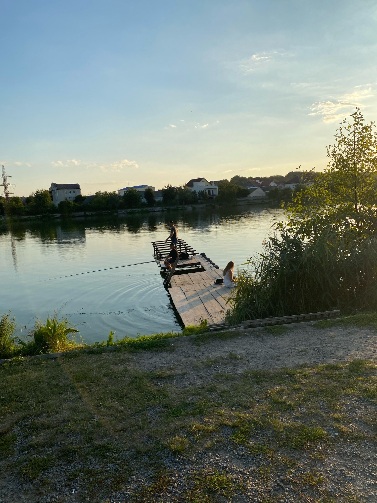 This is a full shot of a wooden pier on a lake in Vinnytsia, Ukraine. The sky is clear blue with a few wispy clouds. The sun is setting, casting a warm golden light on the scene. There are several people on the pier. Two men are fishing, with fishing rods extended into the water. A woman is sitting on the edge of the pier, facing away from the camera. The lake is calm, with gentle ripples on the surface. Trees and houses can be seen on the far bank of the lake. The pier is made of weathered wood and appears to be well-used. The overall atmosphere is peaceful and serene.