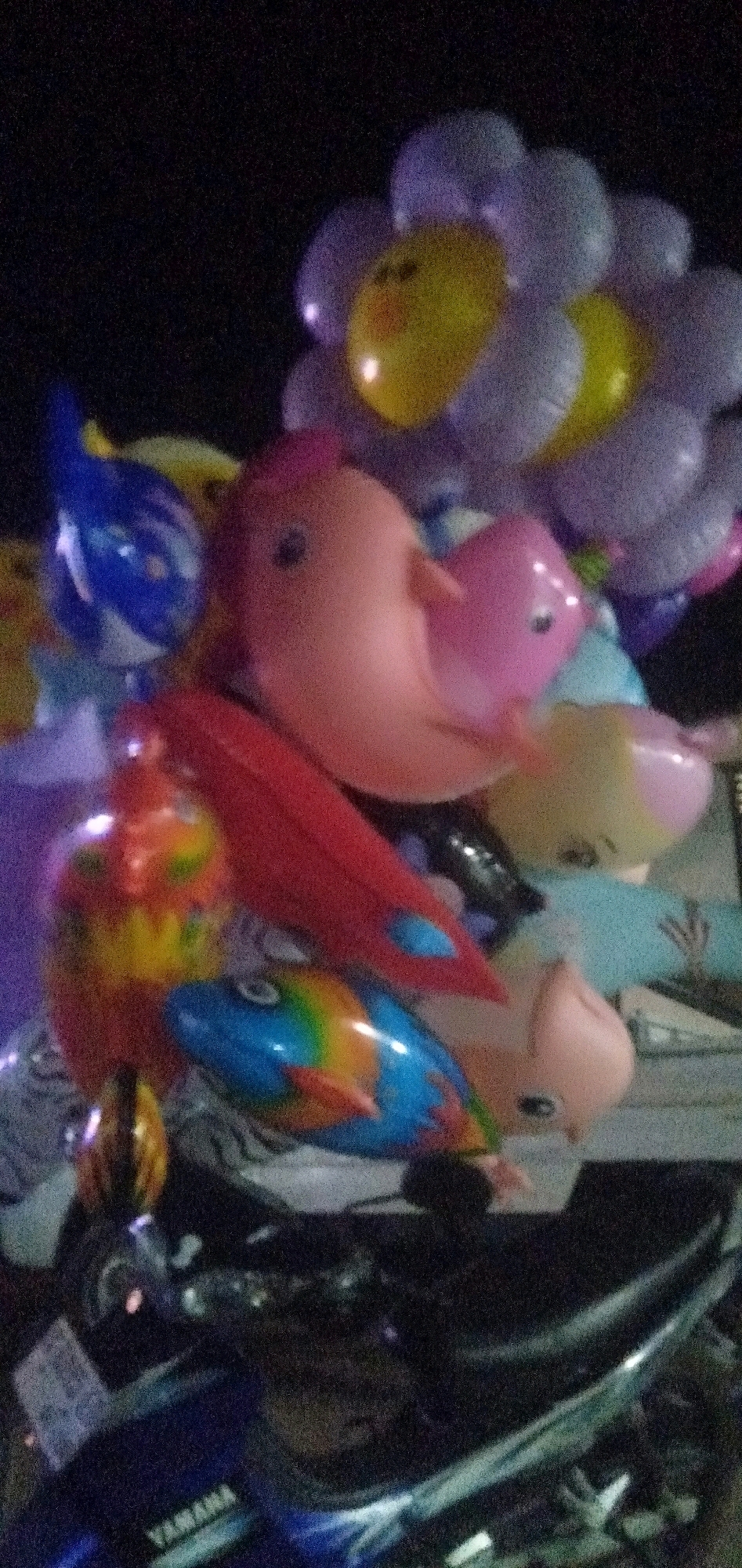 In Plemahan, Indonesia, a collection of colorful inflatable balloons is displayed on a dark background, possibly at night. The balloons are shaped like various characters, including a flower with a yellow center, pink birds, and fish with rainbow patterns. Some of the balloons appear to be lit, giving them a glowing effect. The scene also includes the front portion of a dark-colored motorcycle, with the "YAMAHA" logo visible on its blue bodywork. The overall impression is one of a festive or decorative arrangement, likely set up for sale or display.