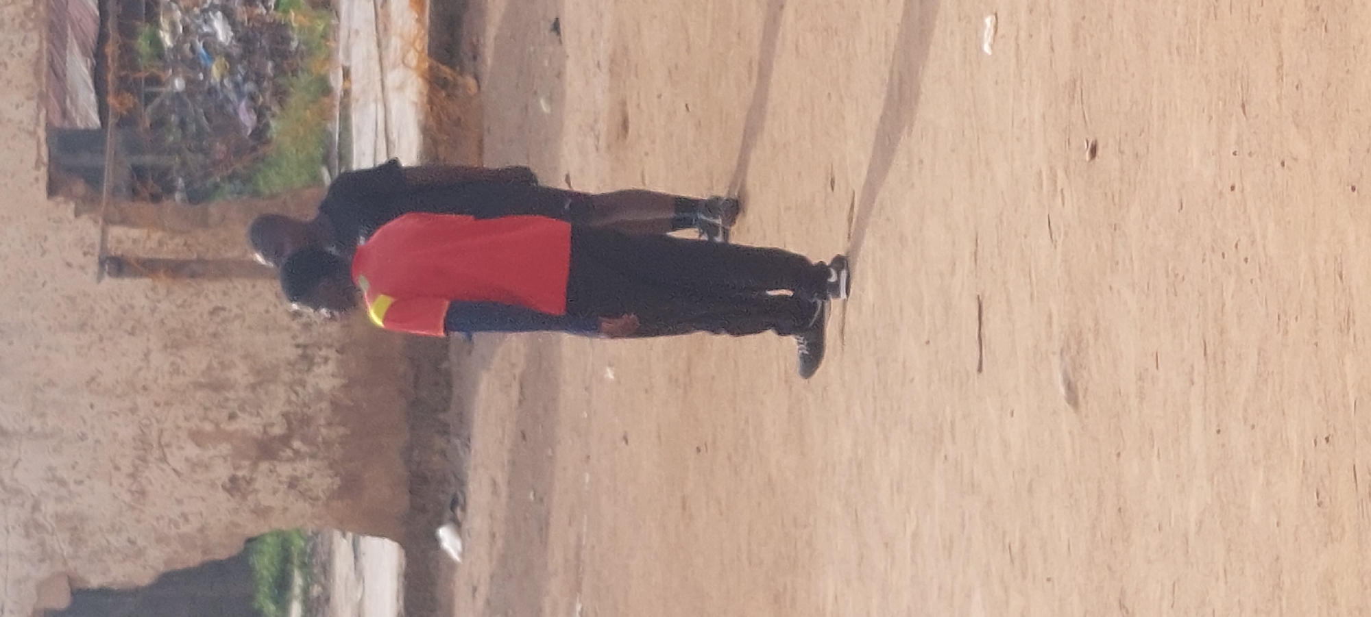 Two individuals are standing on a dirt surface in Bukuru, Nigeria. The person in the foreground is wearing a red shirt with yellow accents on the shoulders and dark pants. This individual is facing away from the camera, with their left side visible. To the right and slightly behind, a second person is wearing a black t-shirt and dark shorts. The background features a damaged, light-colored building with visible textures and some debris piled behind it, suggesting a potentially unkempt urban environment. The ground is dusty and uneven, with shadows cast on the surface.