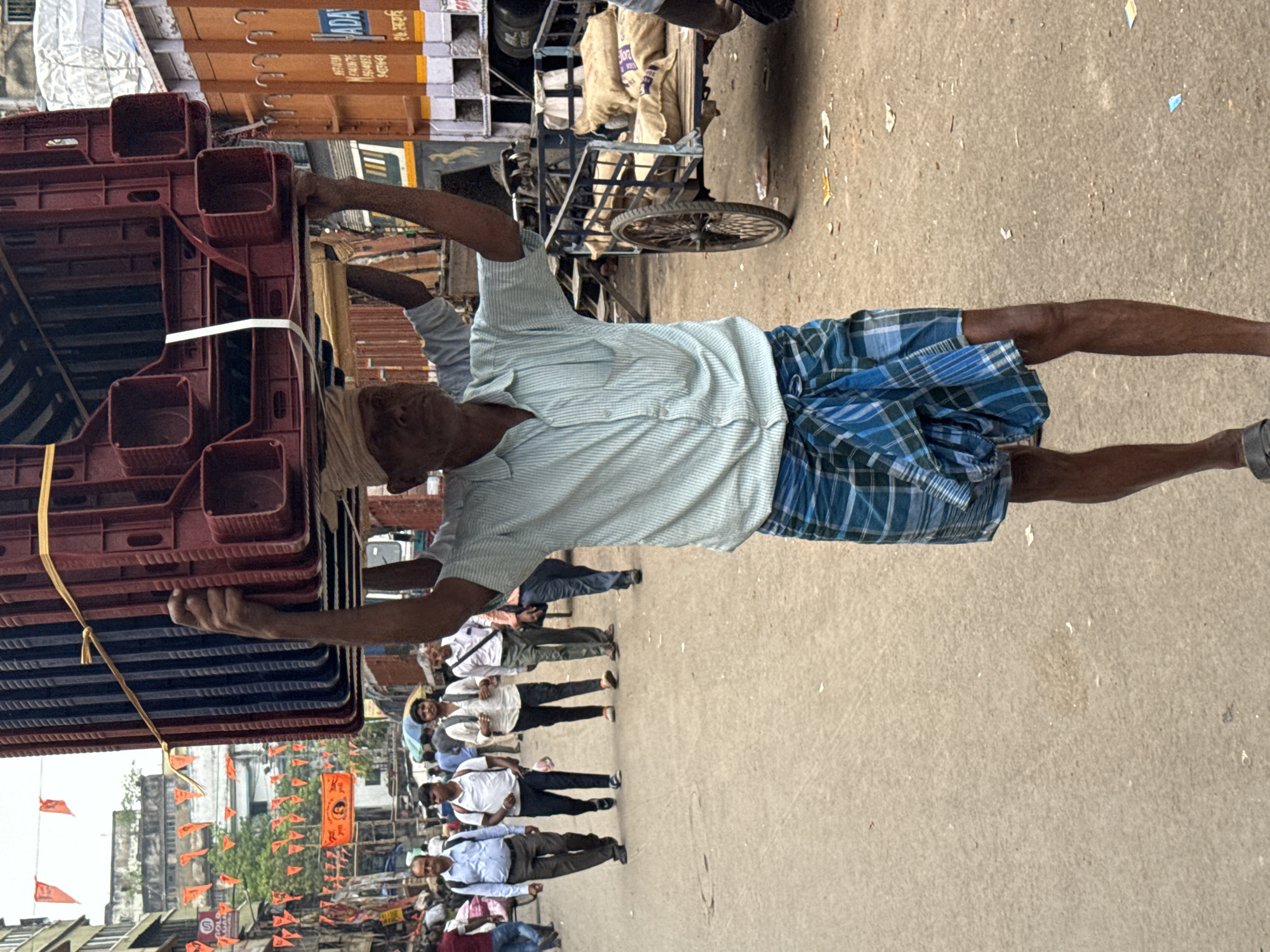 An elderly man in Kolkata, India, is carrying a stack of red crates on his head. He is wearing a light blue striped shirt and a blue and white checkered sarong. A crowd of people, some wearing white shirts, are walking down the street in the background, past a line of orange pennants. A three-wheeled vehicle and a truck are parked nearby.