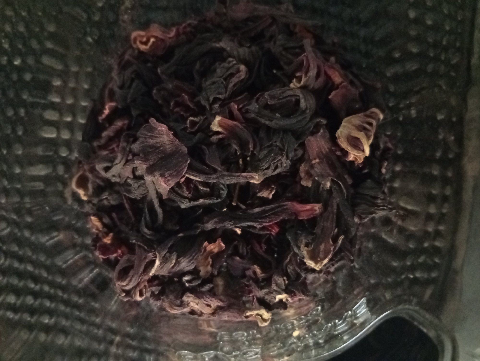 The image shows a pile of dried hibiscus flowers in a glass jar. The flowers are mostly dark red and brown in color, with some lighter yellowish pieces mixed in. The jar appears to be textured on the outside.
The photograph was taken looking down into the jar. There is no evidence to determine if the image was taken in the city of Buenos Aires, Argentina.