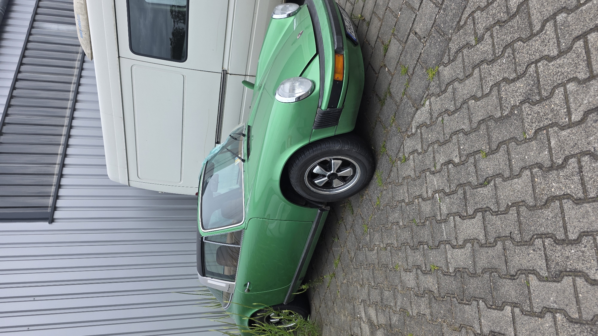 An eye-level, medium outdoor shot shows a vintage green Porsche Targa parked on a grey cobblestone surface in front of a corrugated metal building and a white van. The car is angled slightly towards the viewer, with its front right wheel closer to the camera. The car has a black targa top, brown interior, and silver alloy wheels. The building to the left is light grey and has vertical lines. The white van to the right is partially visible and appears to be a cargo van. The cobblestones are interlocking and have small patches of grass growing between them. The lighting is overcast, casting a soft, even light on the scene.