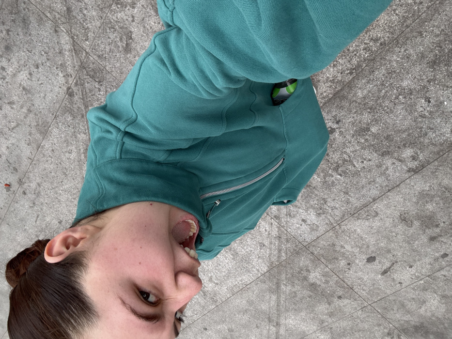 From a high-angle perspective, a person's head and upper torso occupy the left side of the image. The individual, positioned at an unusual angle, is wearing a teal-colored hooded zip-up jacket. Their mouth is open wide, possibly shouting or laughing. Dark hair is pulled back in a bun. The background consists of gray concrete pavement with subtle patterns. A green object is visible in the jacket's pocket. The lighting seems natural and diffused.