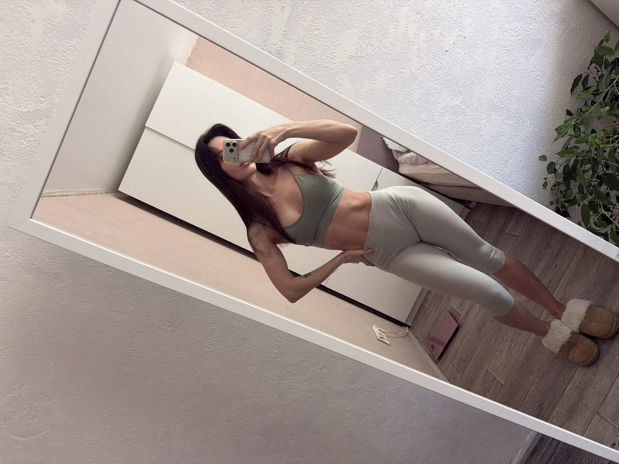 A woman is reflected in a large rectangular mirror, holding a phone in her right hand to take a picture of herself. She's wearing a light green sports bra and matching capri leggings, with tan slippers on her feet. Her hair is long and dark. Behind her, a white dresser is visible, along with parts of a bed. The wall is light in color, with a textured finish. A green leafy plant is in the background on the right. The floor appears to be wood. A scale is visible on the floor. This image was taken in Perm, Russia.