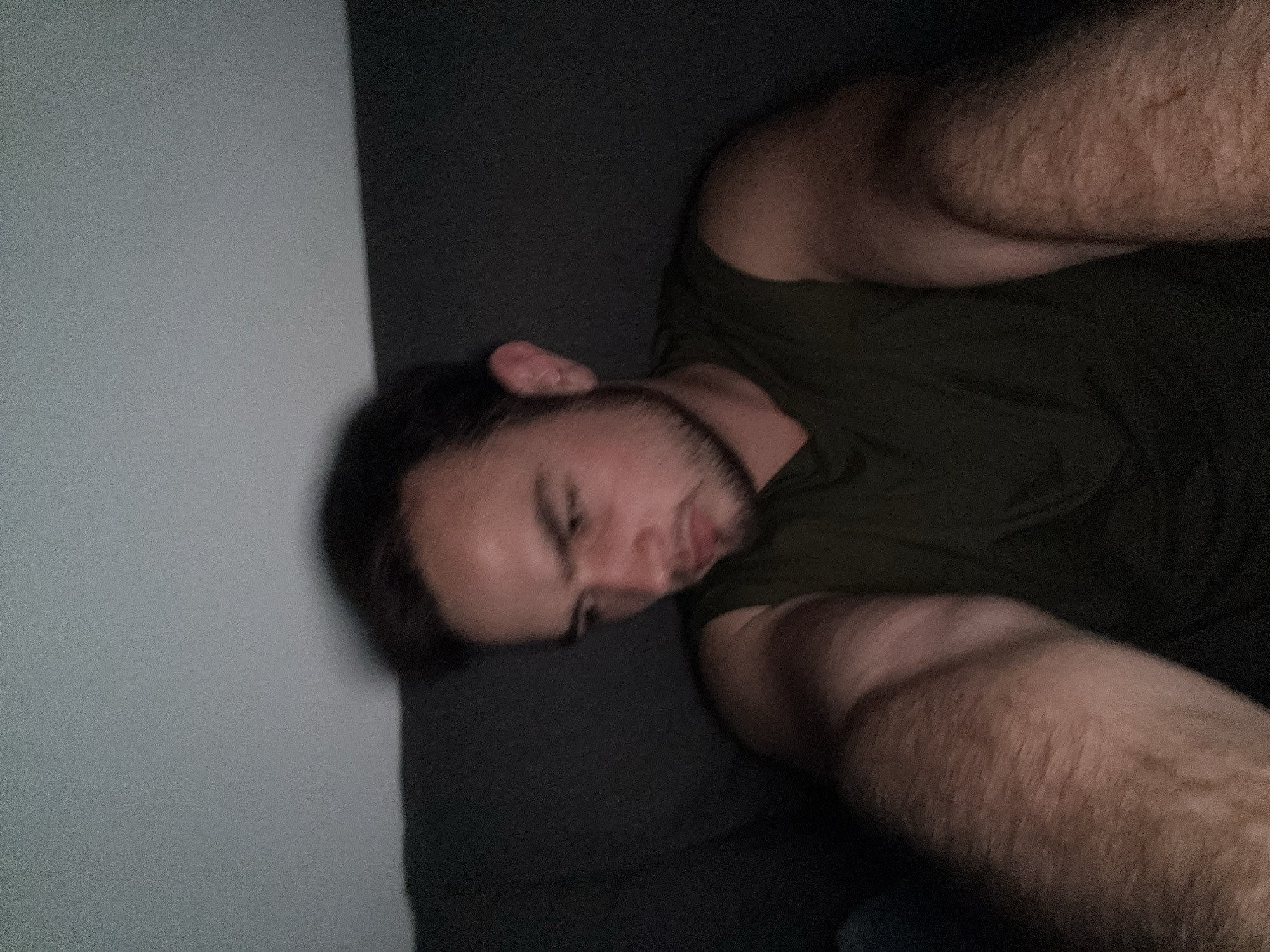 The image shows a man lying down indoors, presumably resting or sleeping. He is positioned on a dark-colored surface, likely a bed or couch, with a plain light-colored wall visible behind him on the left. The man has dark hair and a beard, and is wearing a dark green t-shirt. His muscular, hairy right arm is prominent in the foreground, extending towards the viewer. He is lying on his side, with his face partially visible and eyes closed or downcast.
The lighting in the room is dim, suggesting a nighttime setting or a room with minimal light. The photograph is taken from an elevated, angled perspective, looking down at the man. The overall scene conveys a sense of calm and repose. No visible text is present in the image. The scene is located in San Bernardino Tlaxcalancingo, Mexico.