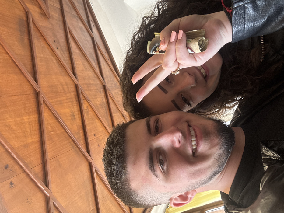 Two individuals, a male and a female, are depicted in a close-up, likely selfie, photograph taken indoors in Kotel, Bulgaria. The male, positioned on the left, has short dark hair with faded sides and a short, trimmed beard. He wears a dark shirt and a dark, possibly synthetic material, jacket, and smiles towards the camera. The female, on the right, has long, dark, curly hair and wears a dark jacket. She also smiles towards the camera, holding her right hand up with the index and middle fingers extended in a 'V' gesture. Her fingernails are painted light pink, and a ring is present on her right ring finger. In her hand, she holds several folded paper currency notes. A red string bracelet is partially visible on her left wrist. The background features a geometrically patterned wooden ceiling composed of light brown panels framed by darker wood strips. A light-colored wall and a yellow curtain are partially visible on the left side of the frame.