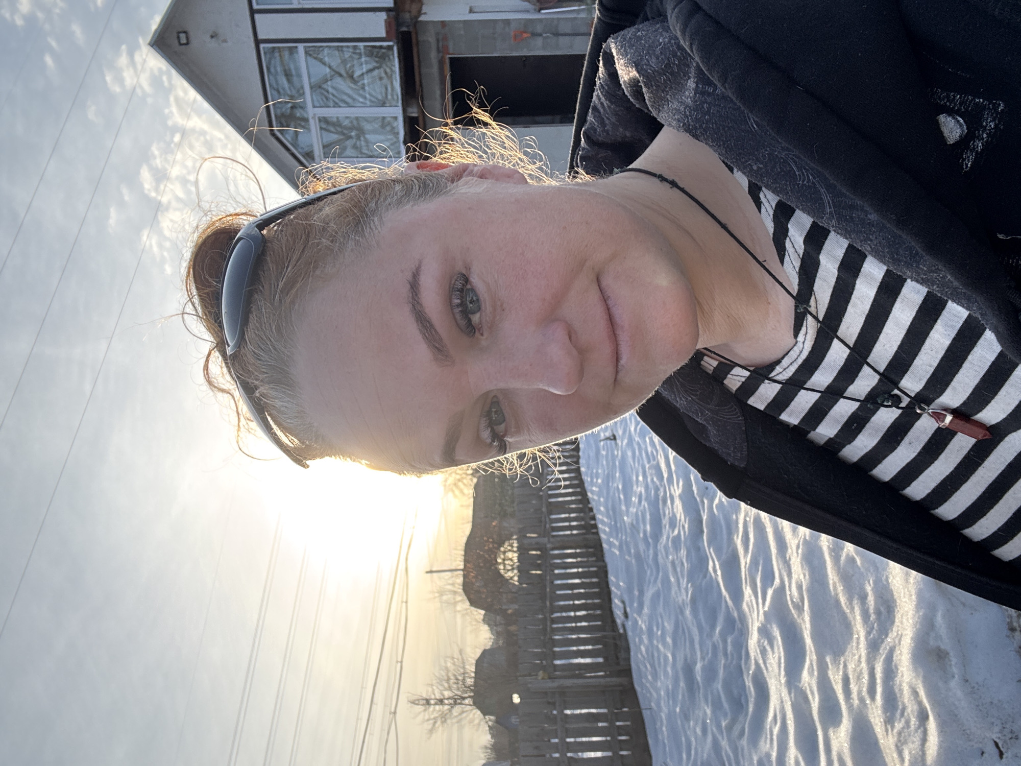 A woman is outdoors in a snowy environment. She is wearing a black and white striped shirt, a black hoodie, and sunglasses perched on her head. She is wearing a necklace with a reddish-brown pendant. The background includes a partially constructed or damaged building, a wooden fence, and trees. The sun is low in the sky, casting a golden glow. Based on the information provided, the location is Chaikovsky, Russia.