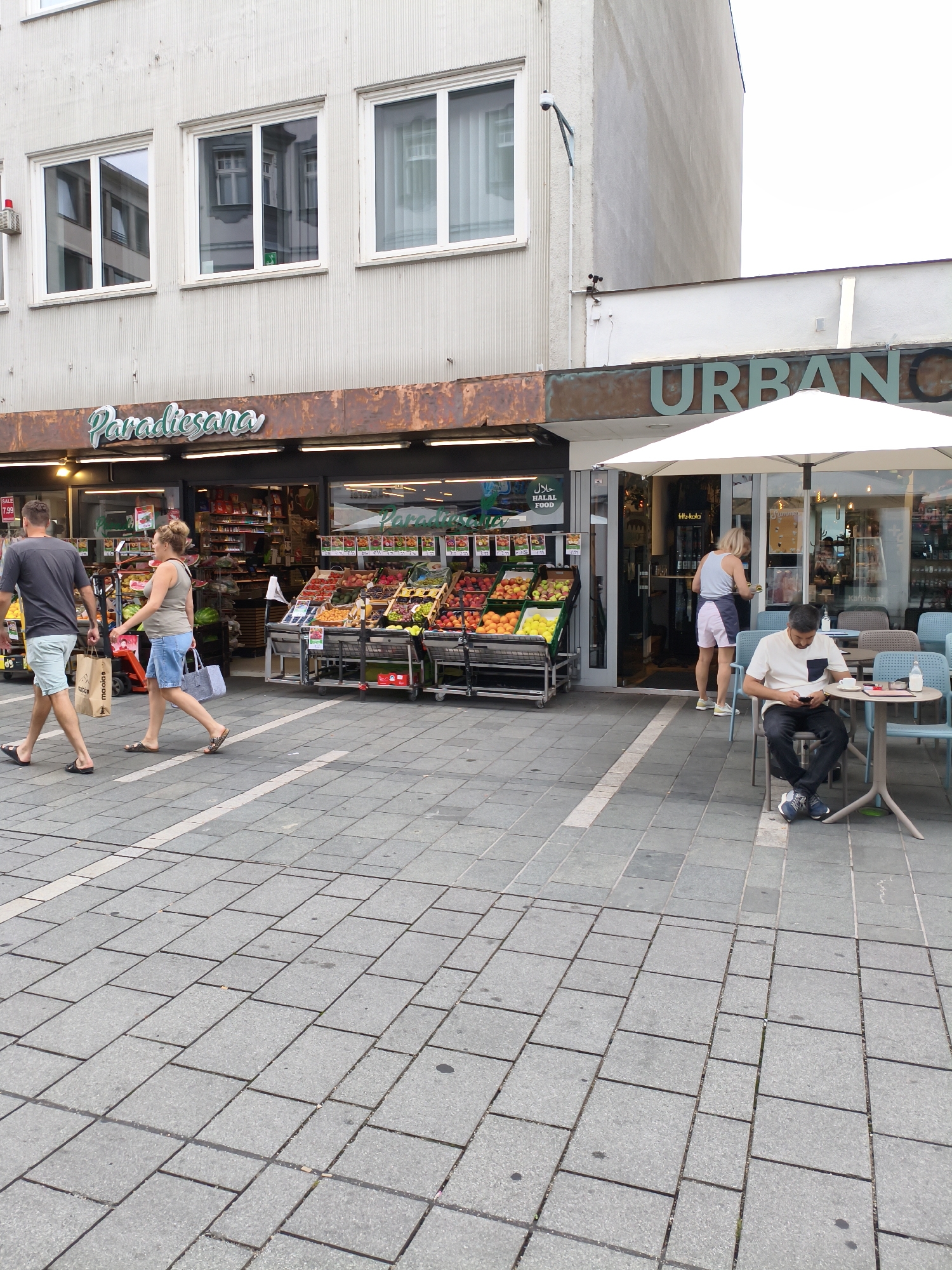 The image depicts a street scene in Regensburg, Germany, featuring storefronts and pedestrian activity. On the left, a store labeled "Paradiesana" displays a colorful array of fruits and vegetables on carts outside its entrance. Two individuals, a man and a woman, are walking past the store, with the man carrying a paper bag and the woman holding a light blue tote bag.
Adjacent to "Paradiesana," another establishment with the prominent sign "URBAN" is visible, featuring an outdoor seating area with a white umbrella. A man is seated at a small table in this area, looking at his mobile phone, with a cup and a bottle of milk on the table. Another person, a woman, stands near the entrance to the "URBAN" establishment. The ground is paved with grey square tiles. The buildings in the background have a concrete facade with several windows. The overall scene suggests a typical urban commercial area.