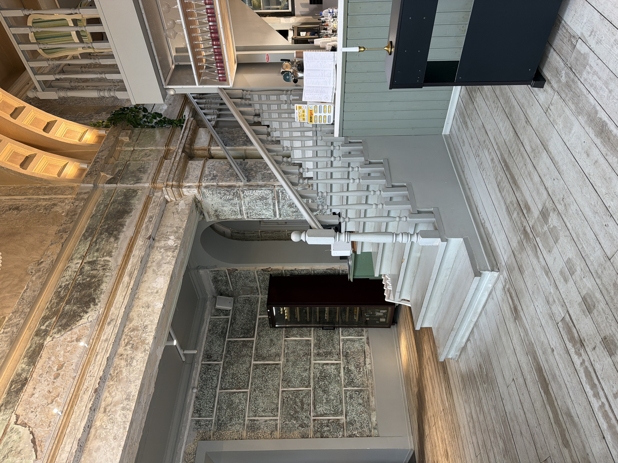 The image shows an interior space with a mix of architectural styles. There's a grand, old-style wall with ornate details and weathered texture, contrasted by a modern-looking staircase with white balusters. The staircase leads to an area with light-colored plank flooring and what appears to be a menu display, near a kitchen or service area. Under the staircase is a wine display. The overall scene suggests a blend of historical and contemporary design elements. It's not possible to determine from the image if this interior is located in Перовское сельское поселение, Ukraine.