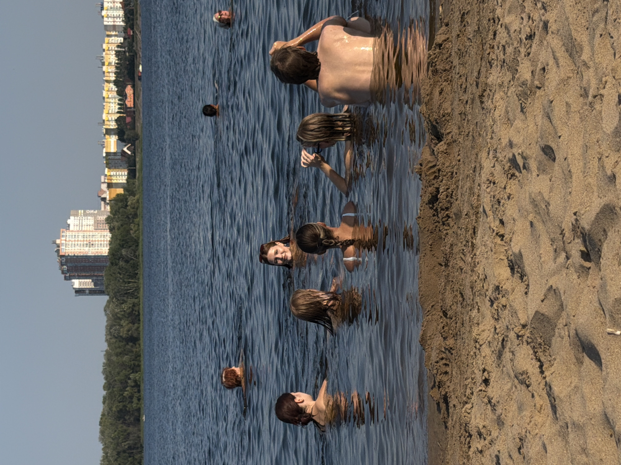 A group of people are swimming in a body of water, likely the Volga River, near the Saratov embankment in Saratov, Russia. The background features a treeline and a cityscape with several tall buildings. In the foreground, a sandy beach is visible. The people are scattered throughout the water, with some closer to the shore and others further out. The lighting suggests it is daytime, possibly late afternoon, with the sun casting a warm glow on the scene. The water is calm with gentle ripples.