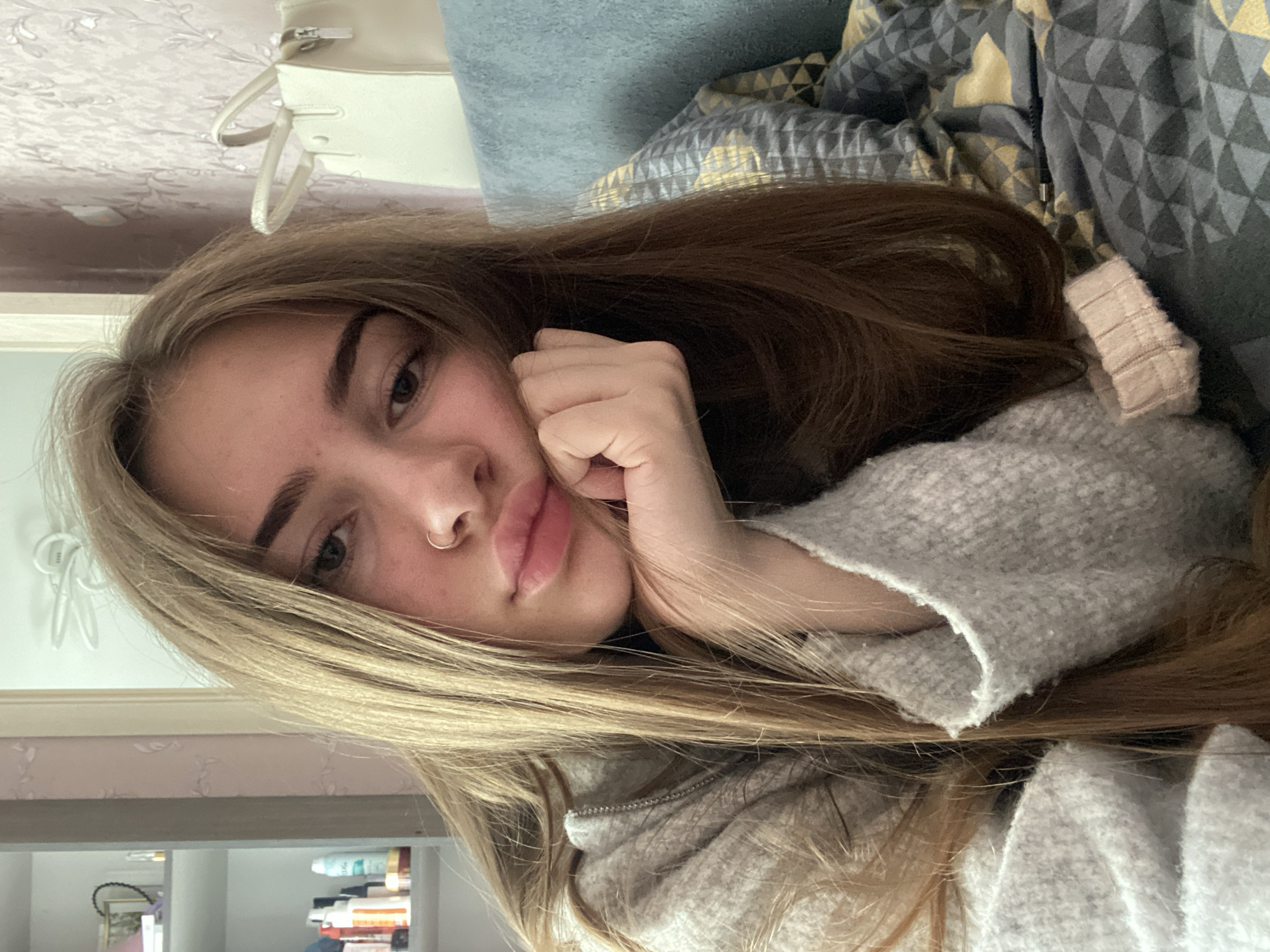 A young woman with long, straight light brown hair is depicted in a close-up, chest-up shot. The image is rotated 90 degrees counter-clockwise, showing her lying on her side, propped up with her left hand supporting her chin. She looks directly at the camera with a neutral to slightly contemplative expression. Her lips are slightly parted, and she has a small nose ring on her left nostril.
She is wearing a light grey, possibly fleece or knitted, hooded jacket or sweater with a visible zipper down the front. Her long hair cascades around her head and shoulders, blending into the textured fabric of her attire.
The background suggests an indoor setting, likely a bedroom or living room. Behind her head, there are soft furnishings, including a plain blue or grey textured pillow/blanket and another patterned one with a geometric grey and gold/yellow design. In the upper left background, a light-colored (cream or off-white) handbag with handles is visible, hanging from an unseen fixture. The lower background (when oriented correctly) shows blurred shelves or furniture with indistinct items, possibly bottles or books. A white cable or wire is also visible on a light-colored wall.
The overall impression is one of a relaxed moment, possibly a casual selfie taken in a personal space. There is no visible text in the image.