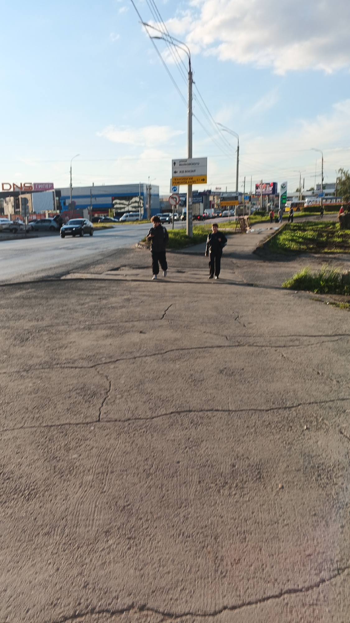 The image depicts an outdoor scene on a paved area, likely a sidewalk or pedestrian crossing, with a street visible in the background. Two young males are walking towards the viewer. The male on the left is wearing a dark hooded jacket and dark pants, while the male on the right is wearing a dark jacket and dark pants. Both appear to be moving at a moderate pace. In the background, a multi-lane road is visible with several cars, including a dark sedan in the process of turning. A variety of buildings and commercial signage are present along the street, suggesting an urban commercial or industrial area. One sign with Cyrillic text indicates "DNS" and "ГИПЕР", likely a store or market. Another sign, a directional arrow, points to "ГОРОД МАРКОВСКОГО" and "Ж/Д ВОКЗАЛ" (railway station), and another points to "ТЕХНИКИ РЕМОНТА" (technical repairs). The sky is partly cloudy with a clear blue sky visible. The lighting suggests it is daytime, possibly late afternoon given the angle of the sunlight. The overall environment appears to be in Izhevsk, Russia, indicated by the Cyrillic signage and the typical urban architecture.