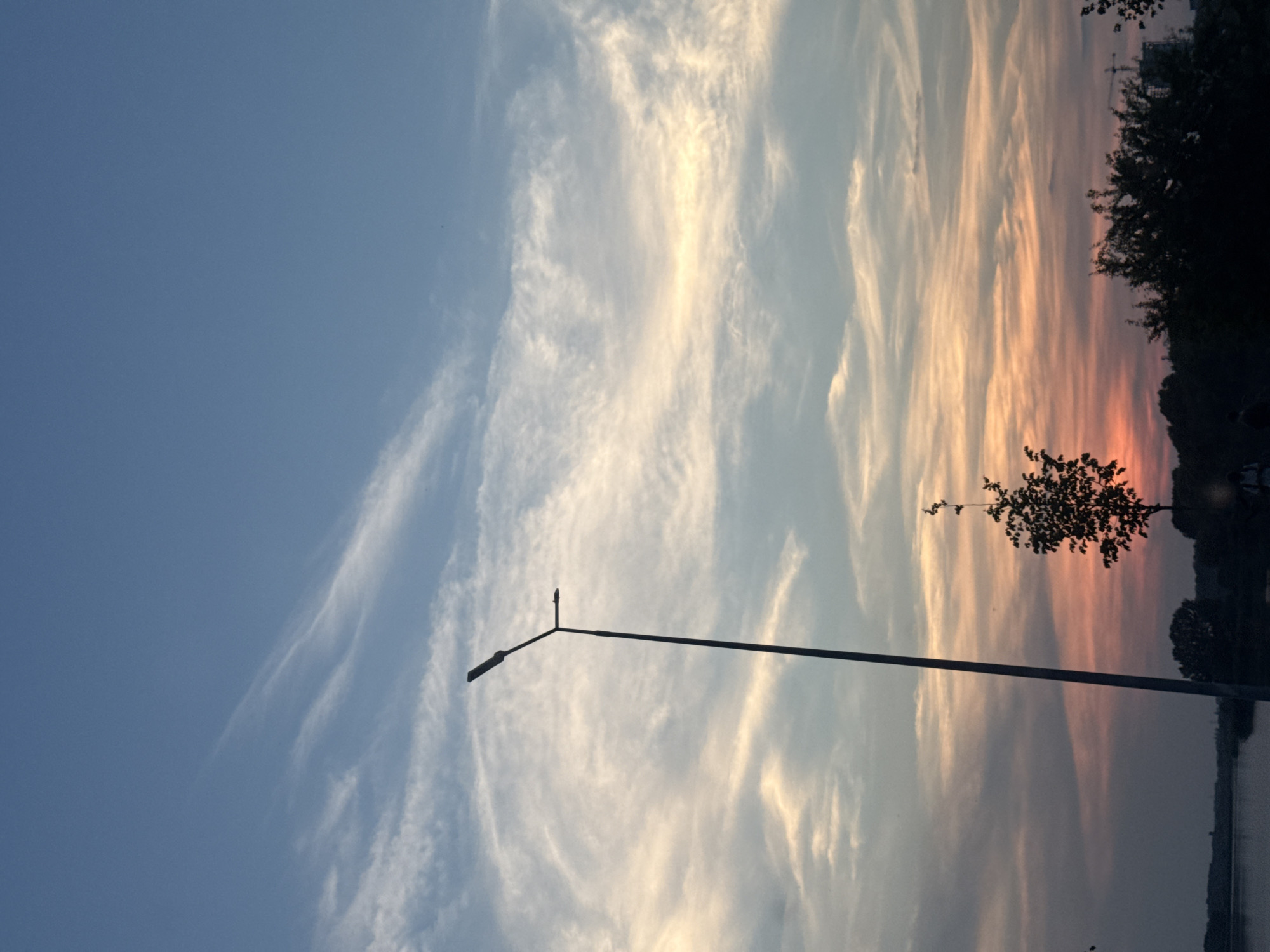 The image captures a sunset over a body of water, likely a river or lake, in Vinnytsia, Ukraine. The sky is filled with wispy, streaky clouds that are illuminated by the setting sun, creating a gradient of colors from a pale blue on the left to shades of orange and pink on the right. A thin, dark silhouette of trees lines the far bank of the water, with hints of buildings and structures visible in the distance. In the foreground, a streetlamp with a distinctive angled arm stands tall against the sky, its dark form contrasting with the vibrant sunset. The water itself appears calm, reflecting the colors of the sky. The overall mood is serene and picturesque, characteristic of a tranquil evening. The time of day is clearly sunset, and the weather appears clear, allowing for a vibrant display of colors. There is no visible text in the image.