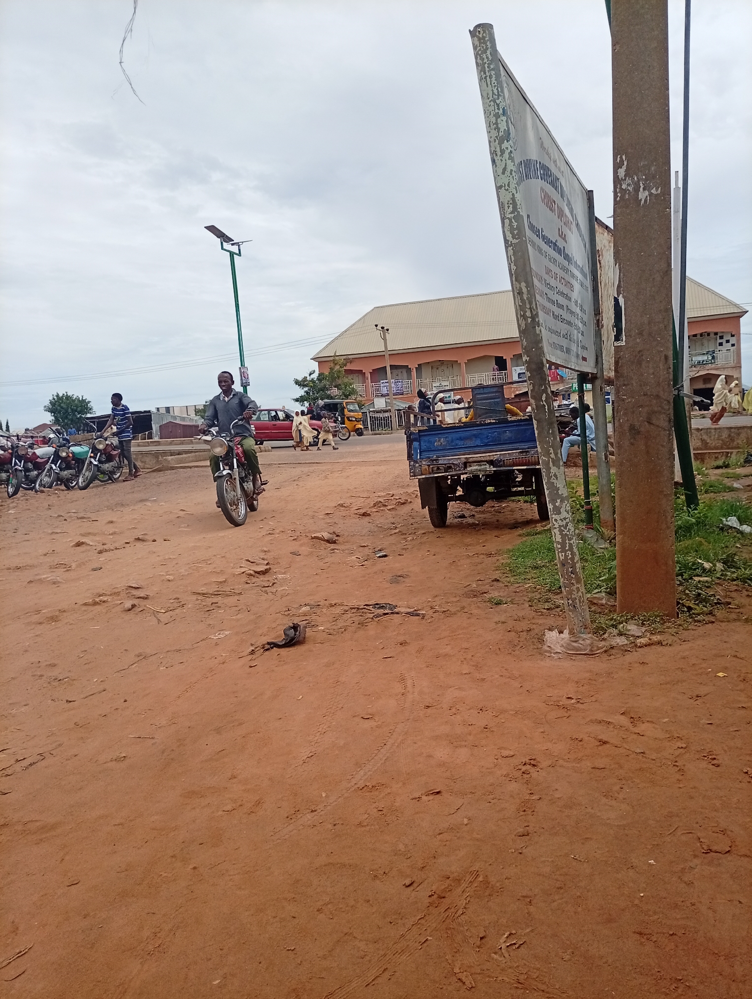In Tumfure, Nigeria, a man on a motorcycle rides on a dirt road under a cloudy sky. To his left, several motorcycles are parked. In the background, buildings line the street, and a blue truck is visible. A large sign stands on a pole to the right of the frame. The ground is covered in red dirt, with tire tracks and debris scattered about.