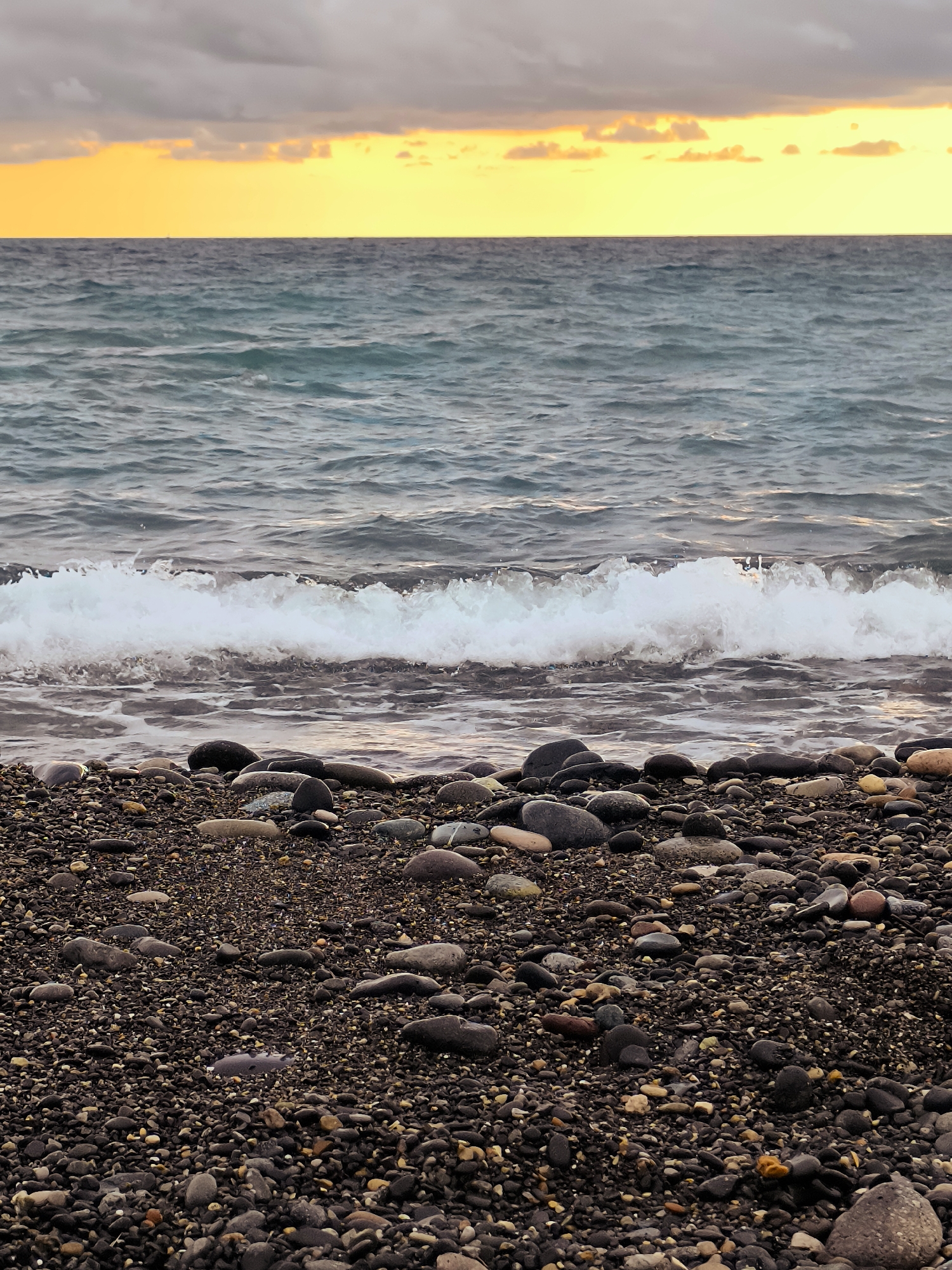 The image depicts a rocky beach scene with the Black Sea in Zemo Kvariati, Georgia. The foreground is dominated by a dense scattering of dark, smooth pebbles and stones, with some larger, rounded rocks interspersed. A gentle wave is breaking on the shore, its white foam contrasting with the darker stones. The water is a deep, muted blue-green, showing ripples from the movement. In the background, a bright, warm yellow band of sky is visible just above the horizon, tinged with orange and gray clouds overhead, suggesting either sunrise or sunset. There are no people or man-made objects visible in the frame.