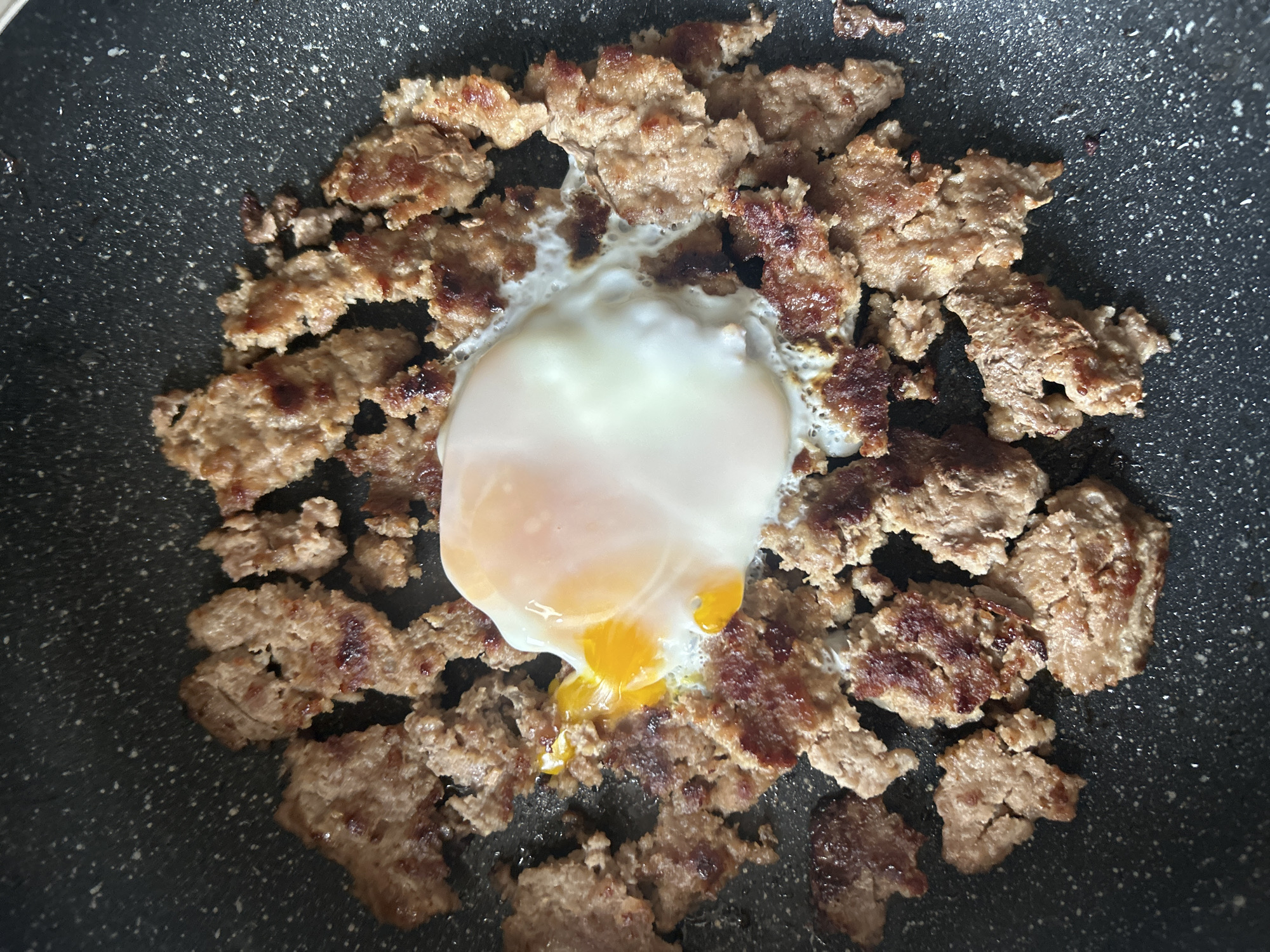 The image shows a close-up of a cooked egg in a frying pan, surrounded by browned, crumbled sausage. The egg appears to be partially cooked, with a runny yolk. The frying pan is black with light-colored speckles. The lighting is bright and the food is in focus. The photograph does not show information relating to Zlatograd, Bulgaria.