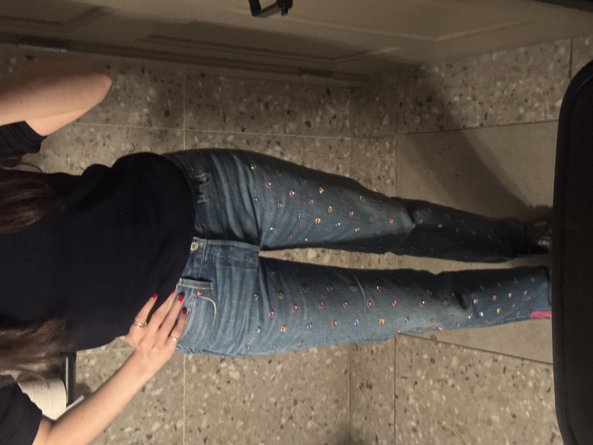 A person wearing blue jeans and a black shirt is lying on a beige, terrazzo-patterned floor. The jeans have colorful embellishments. The person's hands are resting on the jeans near the hip area. The floor appears to be indoors, potentially a home or apartment. The location in the city of New York, United States of America is not possible to determine from the given information.
