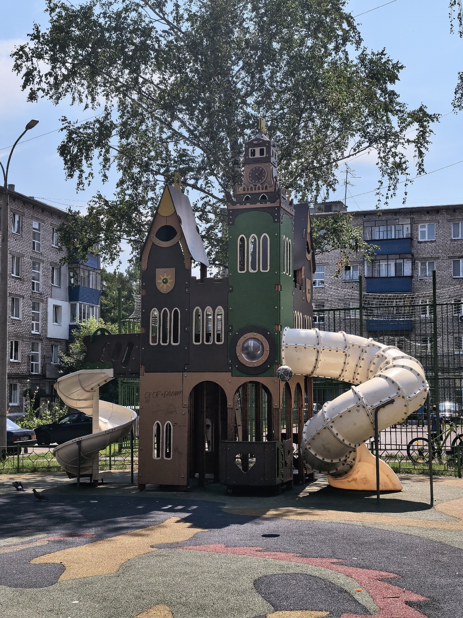 This media file captures a vibrant children's playground in Sterlitamak, Russia, under a bright, sunny sky. The focal point is a large, multi-story play structure designed to resemble a castle, complete with towers, arched windows, and a clock face. Two large, white, spiral tube slides extend from the structure, offering an exciting descent. The ground surface of the playground is a colorful, patterned rubber matting, designed for safety and visual appeal.
In the background, two apartment buildings, constructed with light-colored brick, stand tall, indicating an urban or suburban setting. Trees with full, green foliage are scattered throughout the scene, providing shade and a natural element. A street lamp is visible on the left, and a black metal fence encloses the playground. There is a black car parked on the left side of the image, and a bicycle leans against the fence on the right.
The image appears to be taken during the daytime, given the strong sunlight casting distinct shadows. The weather seems pleasant and clear, suitable for outdoor activities. There are a few pigeons walking on the playground floor. Visible text on the castle structure reads "СГОР-ПАУЛИНН" and "АМ". The overall mood is cheerful and inviting, suggesting a lively atmosphere for children to play and explore.