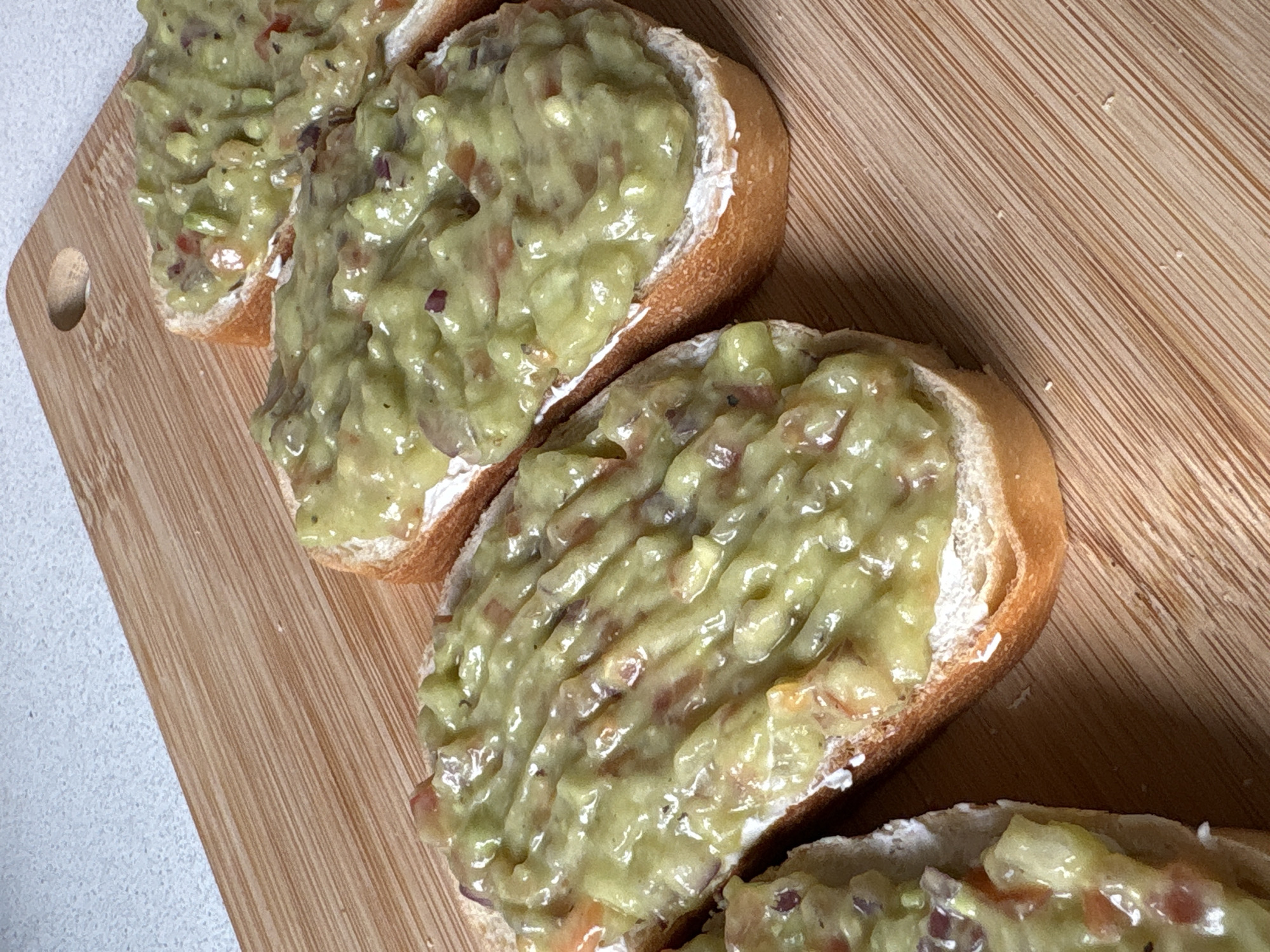The image presents a close-up, overhead view of four appetizer portions laid out on a wooden cutting board. Each portion consists of a slice of baguette-style bread, generously topped with a thick, chunky, light green spread. This spread, likely guacamole, contains discernible pieces of red onion and possibly other diced vegetables, suggesting a fresh preparation.
The bread slices exhibit a golden-brown crust and a softer, lighter interior. They are positioned diagonally across the cutting board, which features distinct vertical wood grain patterns. A small, oval-shaped hole is visible on the left edge of the cutting board.
To the immediate left of the cutting board, a textured, white or light grey surface, presumably a kitchen countertop, is visible. The lighting is bright and even, illuminating the details and textures of the food. The scene depicts a casual food preparation or serving setting, possibly in Saint Petersburg, Russia, though no specific geographical cues are visible in the image itself. There are no people or visible text in the frame.
