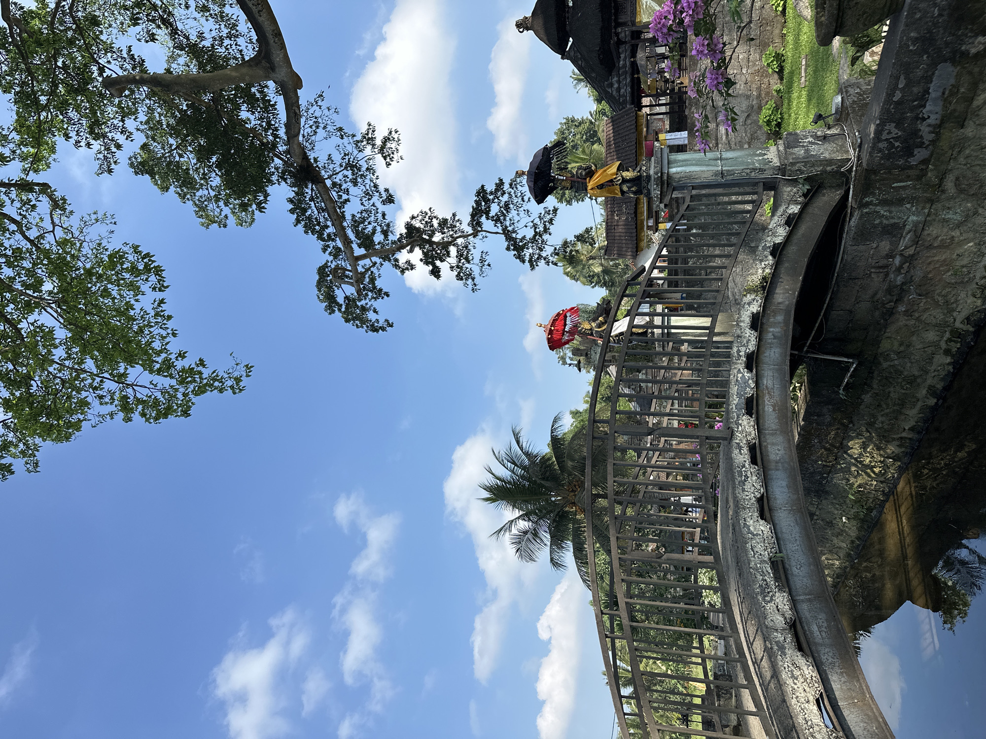 The image depicts a bridge in the city of Marga, Indonesia. The bridge, constructed of metal and stone, spans a body of water, the reflections of trees and the sky visible on its surface. The bridge is decorated with two statues, one holding a red umbrella and the other a black umbrella. Both statues are draped in yellow fabric. Behind the bridge, traditional Indonesian architecture with thatched roofs can be seen. Lush green vegetation, including palm trees, surrounds the area, under a clear blue sky with scattered white clouds. The overall scene suggests a peaceful, cultural setting.