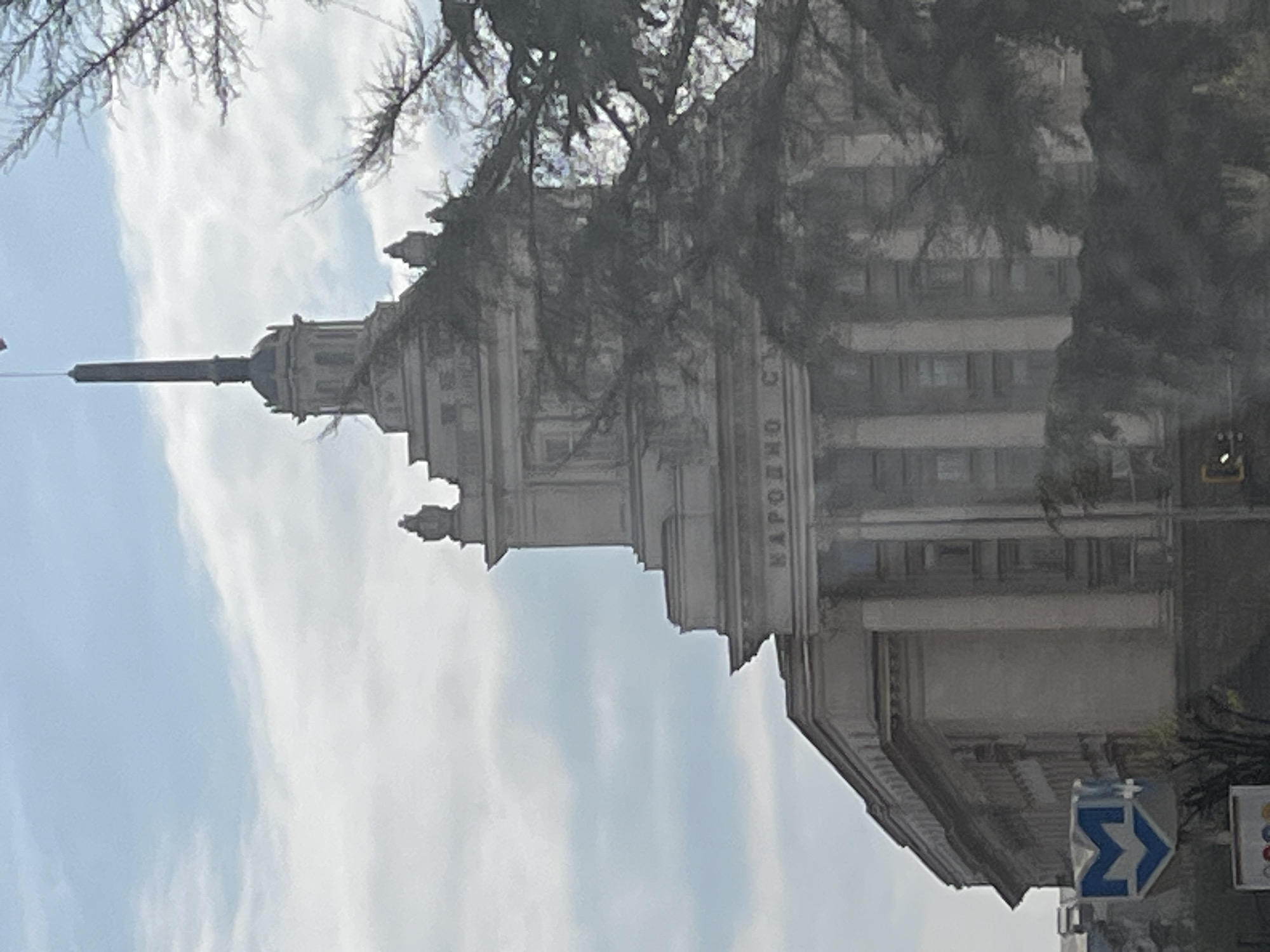 The image, correctly oriented, displays a grand, classical-style building under a partly cloudy, light blue sky. The multi-story structure features a layered design, prominent columns, and ornate details, culminating in a tall, slender spire. Bare tree branches are visible in the foreground, partially obscuring parts of the building.
On one of the building's facades, Cyrillic text is visible, reading "НАРОДНО СЪ". This translates to "National" followed by the beginning of another word, strongly suggesting "National Assembly" or "National Council" in Bulgarian.
In the lower right corner of the image, a blue, arrow-shaped sign with a white stylized "M" or Greek letter sigma (Σ) is present. This symbol is commonly used to indicate a metro (subway) station entrance.
The scene depicts an urban environment during daytime. The weather appears clear with visible clouds. No people or specific activities are evident. The architecture, language, and metro sign strongly suggest a location in Eastern Europe, likely Bulgaria, possibly Sofia.