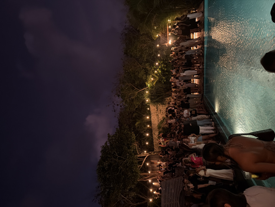 This nighttime image captures an outdoor social event at a venue in Uluwatu, Indonesia. Approximately several hundred individuals, primarily adults, are gathered on a wooden deck bordering a rectangular swimming pool. Attendees are standing, conversing, and many are holding beverages. Their attire varies from casual to semi-formal evening wear. The scene is illuminated by numerous string lights strung between lush trees and overhead structures, casting a warm glow on the crowd and reflecting off the rippled surface of the swimming pool. The background features dense foliage against a dark night sky with visible cloud formations. A textured wall, possibly stone, is evident behind a section of the crowd on the left. A small, illuminated, open-sided pavilion-like structure is visible near the pool on the right. In the immediate foreground, an individual with braided hair, observed from behind, holds a glass containing a light-colored beverage.