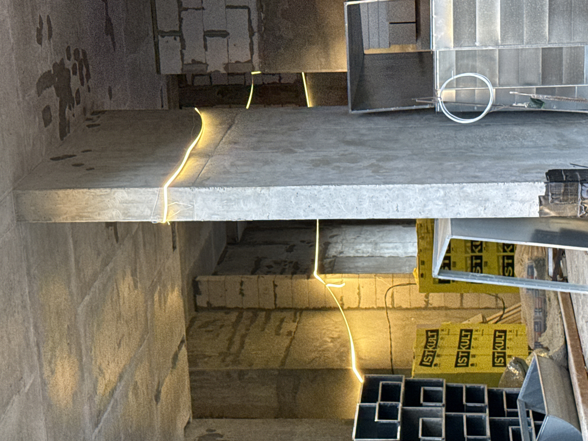 This is an indoor construction site in Moscow, Russia, likely a building under renovation or construction. The scene is dimly lit, with yellow LED strip lights illuminating the concrete walls, ceiling, and columns. On the left side of the image, stacked black metal shelving units are visible. To the right of the shelving, there are several yellow packages with the text "ISTKULT" printed on them. Further into the image, more construction materials, including what appear to be concrete blocks and metal structures, are present. A large concrete pillar runs vertically through the center of the frame, dividing the space. The floor is covered with debris and dust, indicative of ongoing work. There are no people visible in the image.