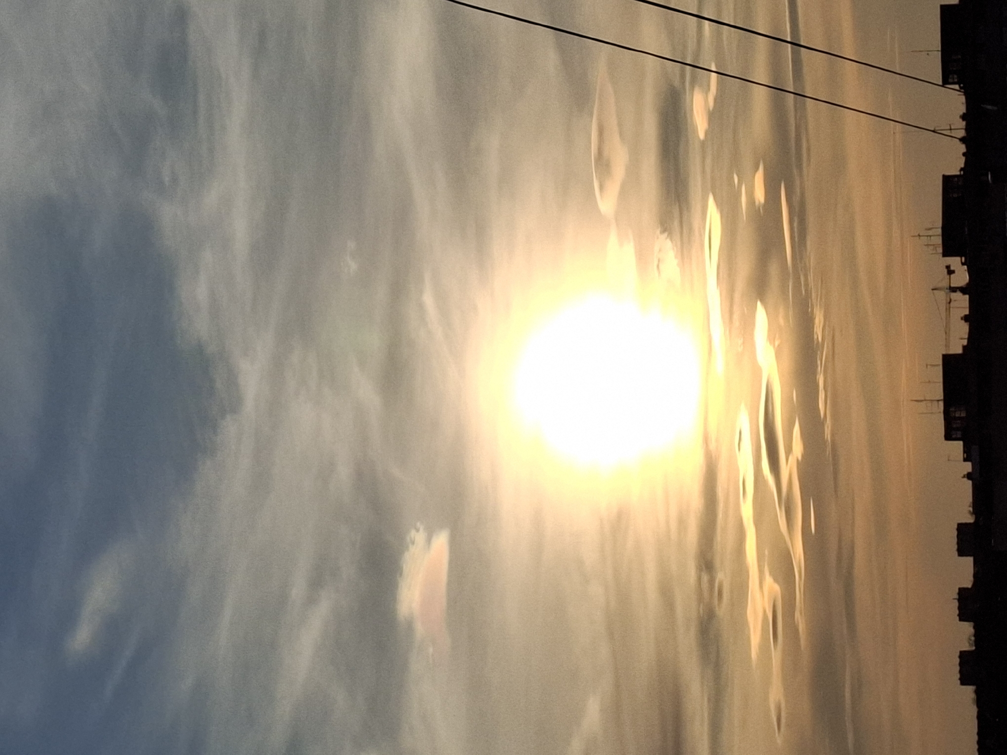 The image shows a bright sun in the sky, possibly at sunset or sunrise. The sky has clouds with varying shades of white and blue. In the background, there's a silhouette of a building skyline. Several wires or power lines are visible running across the upper portion of the frame. Based on the architecture and sky appearance, the location appears to be Novi Sad, Serbia. The strong sun suggests the photo was taken either early in the morning or late in the afternoon.