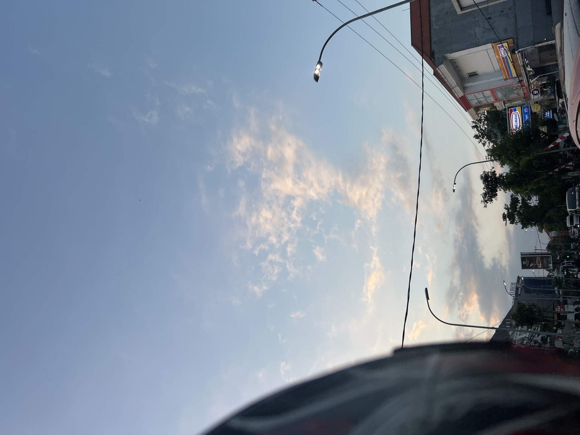 The image captures a street scene in Kuta, Indonesia, at dusk. The sky is a soft blue with scattered wispy clouds tinged with pink and orange, indicating the setting sun. In the foreground, a portion of a motorcycle helmet is visible on the left, suggesting the image was taken from the perspective of a rider. Streetlights with modern LED fixtures illuminate the roadside, casting a warm glow.
Along the street, several buildings are visible. One building on the right has a red-tiled roof and a gray facade, with windows and balconies. A sign for "Indomaret," a popular convenience store chain in Indonesia, is prominently displayed on the building. Other signs and advertisements can be seen on various shops and establishments, hinting at a commercial area.
Trees with lush green foliage line parts of the street, providing a natural element amidst the urban landscape. In the background, more buildings and streetlights are visible, extending into the distance. Traffic appears to be present, with vehicles on the road, although not in heavy congestion. The overall atmosphere is one of a typical evening in a bustling Indonesian city, with the blend of urban infrastructure and the beauty of the twilight sky.