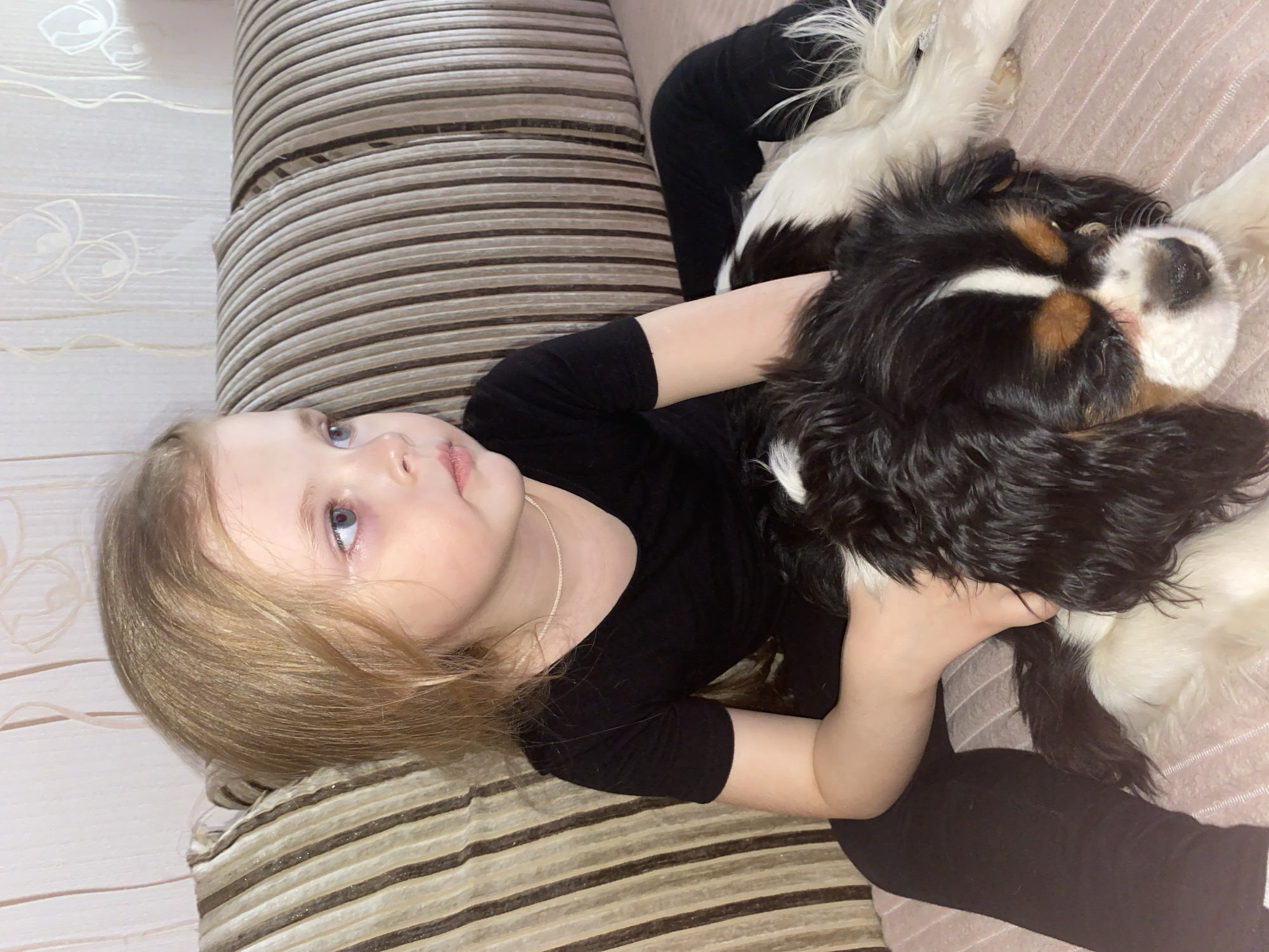 An indoor scene depicts a young child and a dog sharing a moment of quiet companionship. A fair-haired child with blue eyes lies on their back, looking upwards with a soft expression. They are dressed in a black long-sleeved top and black pants. Their left arm is wrapped gently around a medium-sized dog, which lies beside them, also resting. The dog is a tri-color breed, likely a Cavalier King Charles Spaniel, with black, white, and tan markings, and appears relaxed, possibly asleep or very calm.
Both subjects are situated on a plush, striped surface, characteristic of a sofa or large cushion, featuring horizontal bands of brown and beige. Adjacent to the child, a light-colored wall with subtle white floral or abstract patterns is visible. The lighting is soft and even, suggesting an interior setting during the day or under warm artificial light. The overall mood of the image is peaceful and affectionate, highlighting the bond between the child and their pet. No visible text is present in the image. The location context for this cozy domestic scene is Votkinsk, Russia.
