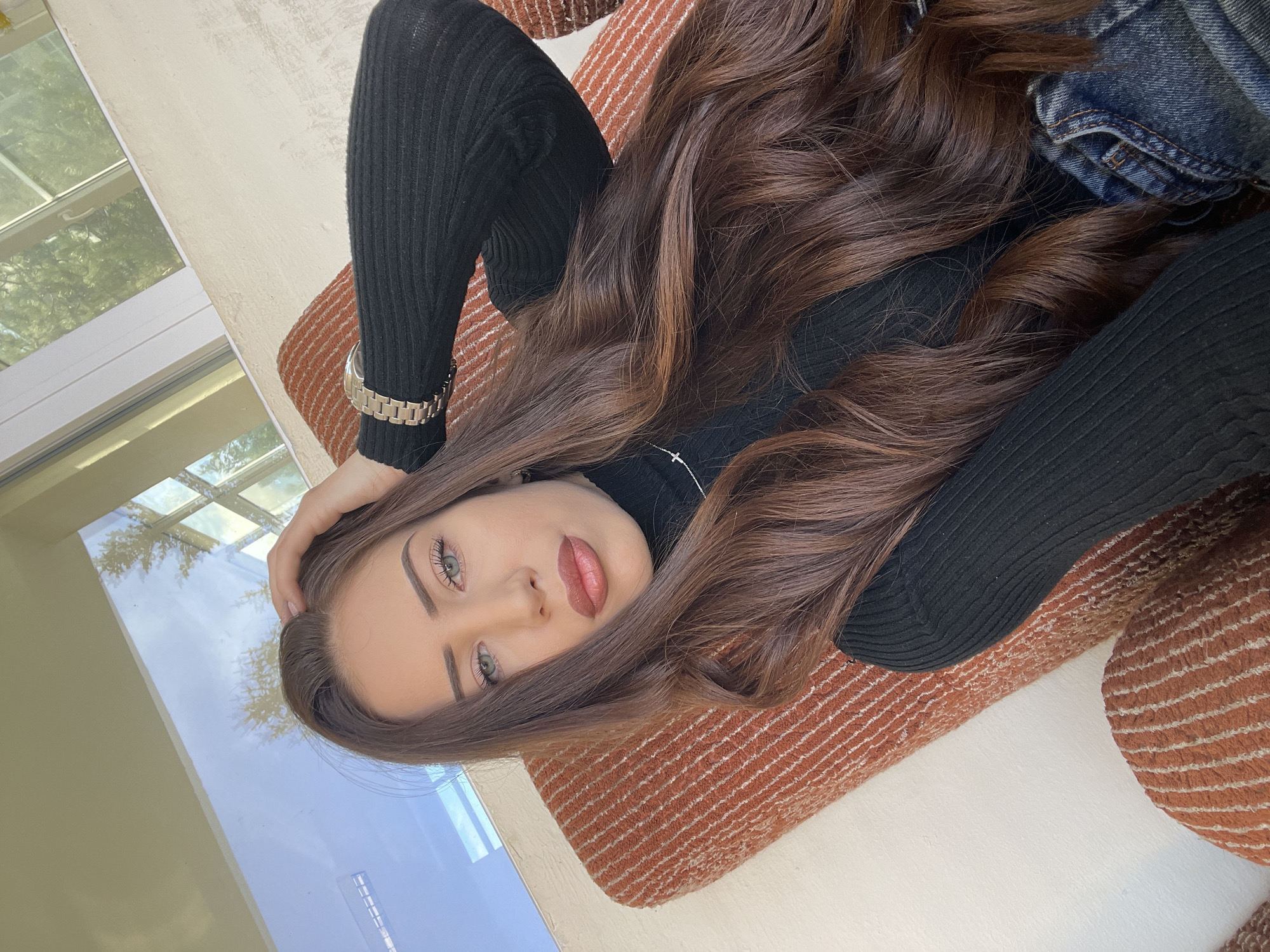 A young woman with light skin, long brown hair, and green eyes is lying down and looking at the camera. She is wearing a black turtleneck sweater and blue jeans. Her hair is styled in loose waves and is draped around her face and shoulders. She is wearing a silver watch on her left wrist. She is resting her left hand on her forehead. She is lying on a textured orange cushion or rug. A window with multiple panes is visible in the background, reflecting the sky and trees. The scene appears to be indoors, possibly in a sunroom or near a large window. Location is unspecified and undeterminable.