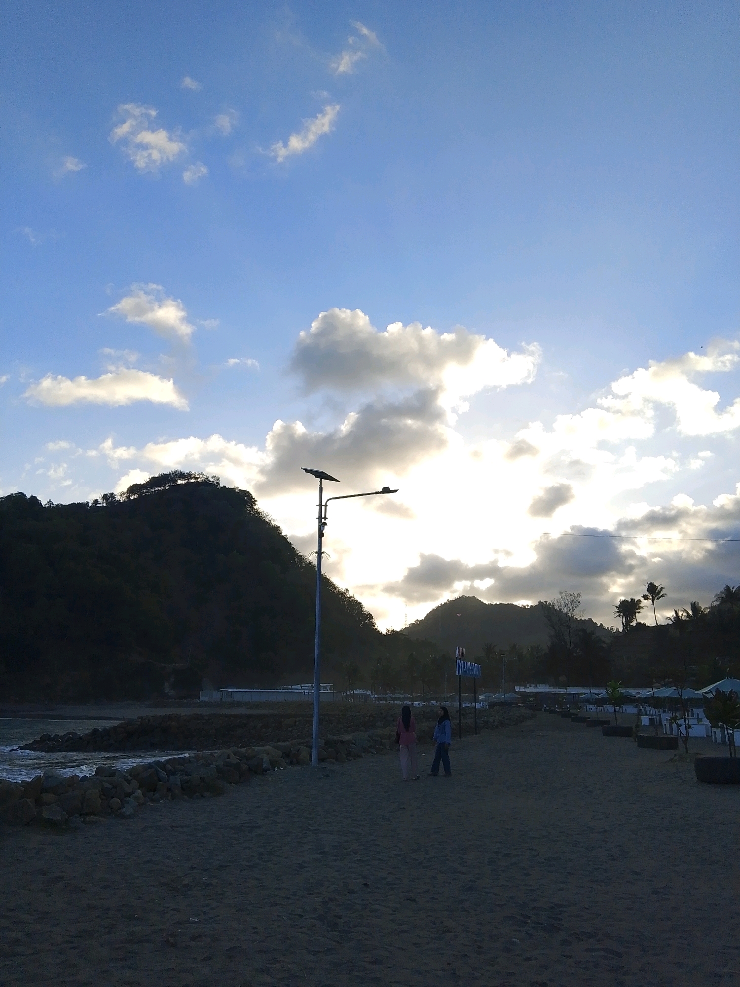 The image depicts a beach scene at Karangbolong, Indonesia. The sky is partly cloudy with the sun setting or rising, casting a bright glow through the clouds. In the foreground, a sandy beach stretches towards the water. A stone embankment lines the edge of the water, with waves gently lapping against it.
Further inland, a paved path runs parallel to the beach. Two individuals, a woman in a pink top and light-colored pants and another person in a blue jacket and dark pants, are walking on this path. A tall lamp post with a solar panel is visible near the path.
In the background, a dark, tree-covered hill or mountain rises behind the beach. Palm trees and other vegetation are scattered along the coastline. Several white structures, possibly beachside facilities or buildings, are also present. The overall impression is of a tranquil coastal environment during twilight.