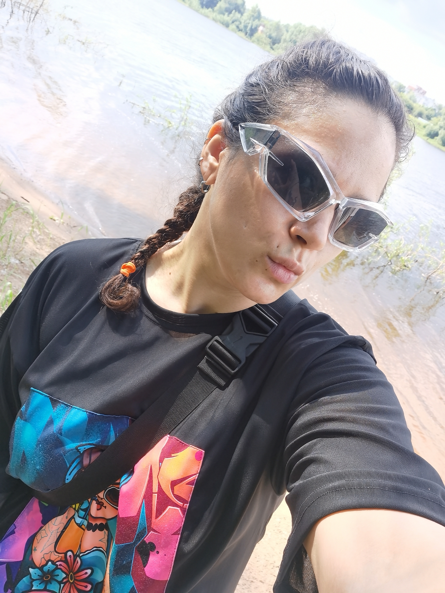 A woman is taking a selfie on the bank of the Kama River in Chaikovsky, Russia. She is wearing a black t-shirt with colorful graffiti-style graphics on the front. She has her dark hair in two braids, with an orange elastic band around one of them. She is wearing large, transparent, angular sunglasses. The river is visible in the background, with trees and buildings on the opposite bank. The area appears to be a sandy or muddy bank.