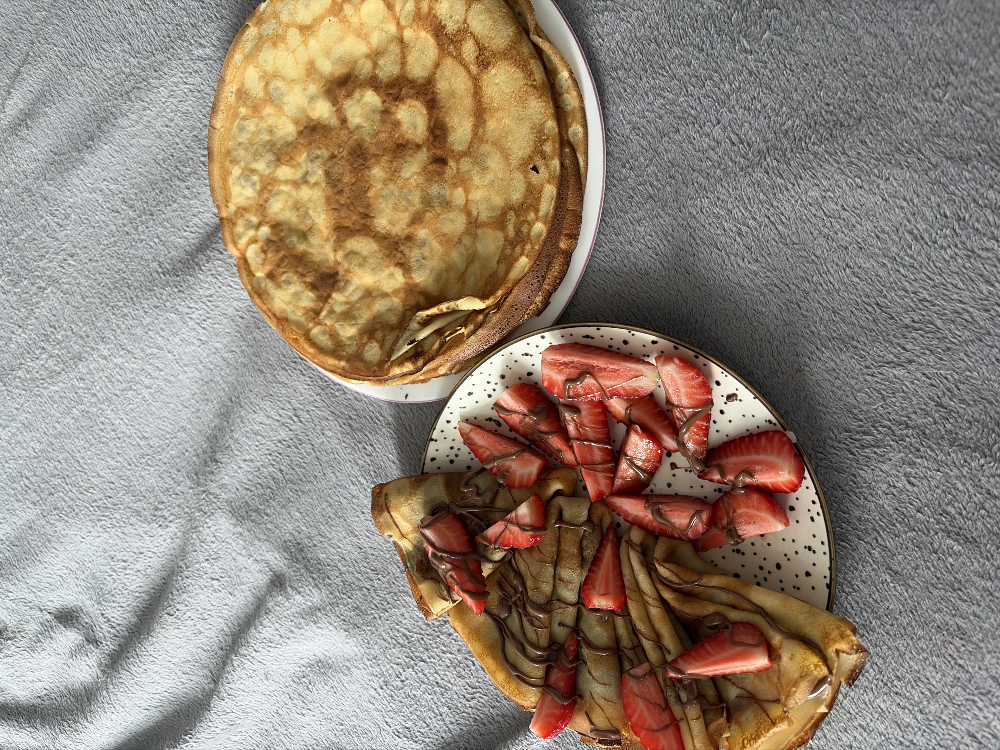 The image shows two plates of food placed on a grey fuzzy surface. One plate holds a stack of several golden-brown pancakes. The second plate contains folded pancakes topped with sliced strawberries and drizzled with chocolate sauce. The location is Новинки, Russia.