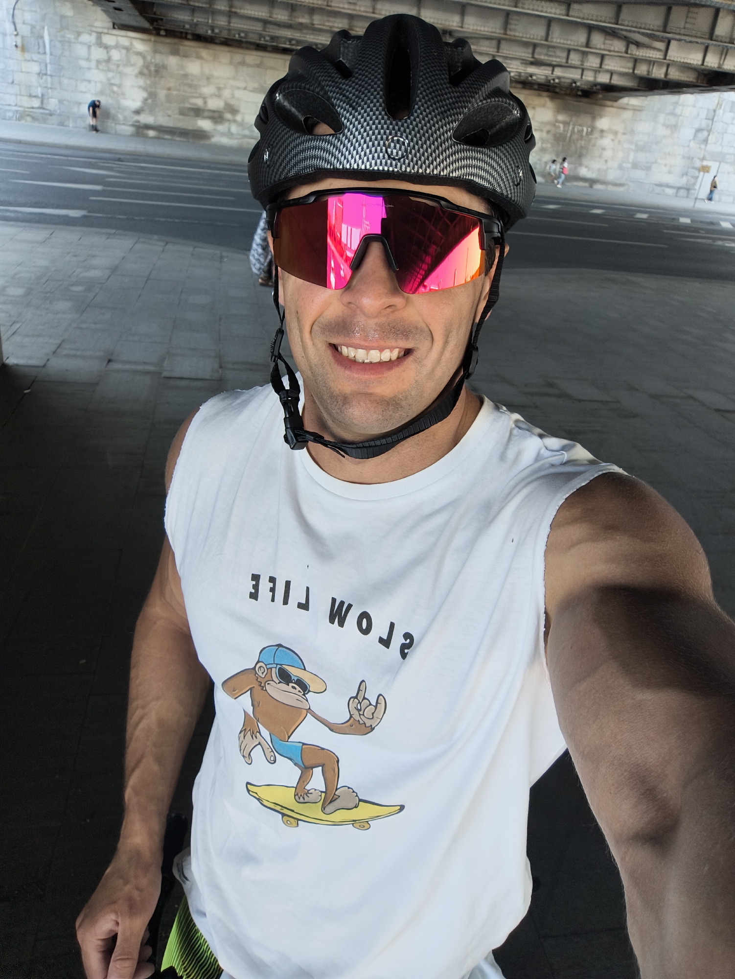 The image captures a selfie of a smiling adult male, positioned within an urban underpass in Moscow, Russia. He is wearing a black cycling helmet with a carbon fiber pattern and pink mirrored sports sunglasses. His white, sleeveless t-shirt, appearing reversed, features a graphic of a monkey in a blue cap and swim trunks on a yellow skateboard, making a 'rock-on' hand gesture, with the text 'SLOW LIFE' visible in reversed lettering above. The man's right arm and hand are partially visible, holding the camera. The background consists of a grey concrete wall on the left, a paved road or pathway with multiple faint white markings, and a metallic bridge structure overhead with visible beams and rivets. In the distance, several small, indistinct figures are visible: one person is bent over on the far left, and multiple individuals are walking further along the underpass on the right.