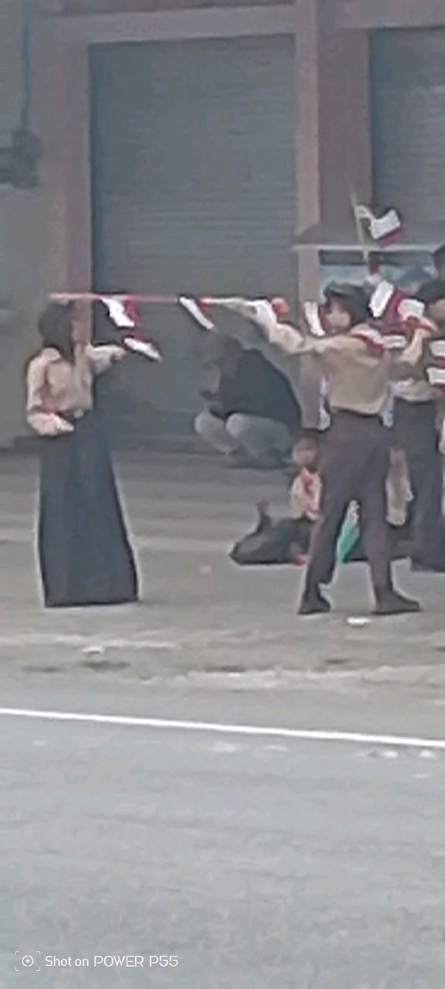 Two individuals, possibly scouts, are holding flags in what appears to be an outdoor public space in Secang, Indonesia. One person on the left, wearing a light-colored shirt and a dark skirt or long dress, holds a flag with red and white stripes horizontally. Another individual, to the right, in a scout-like uniform with a brown shirt and dark pants, also holds a flag with red and white stripes. In the background, a large, gray, corrugated metal door or wall is visible. Several other people are present in the mid-ground, some are crouching or sitting, and another individual is standing. The ground appears to be a paved surface, possibly asphalt, with some white markings. A watermark at the bottom indicates the photo was taken with a "POWER P55" device.
