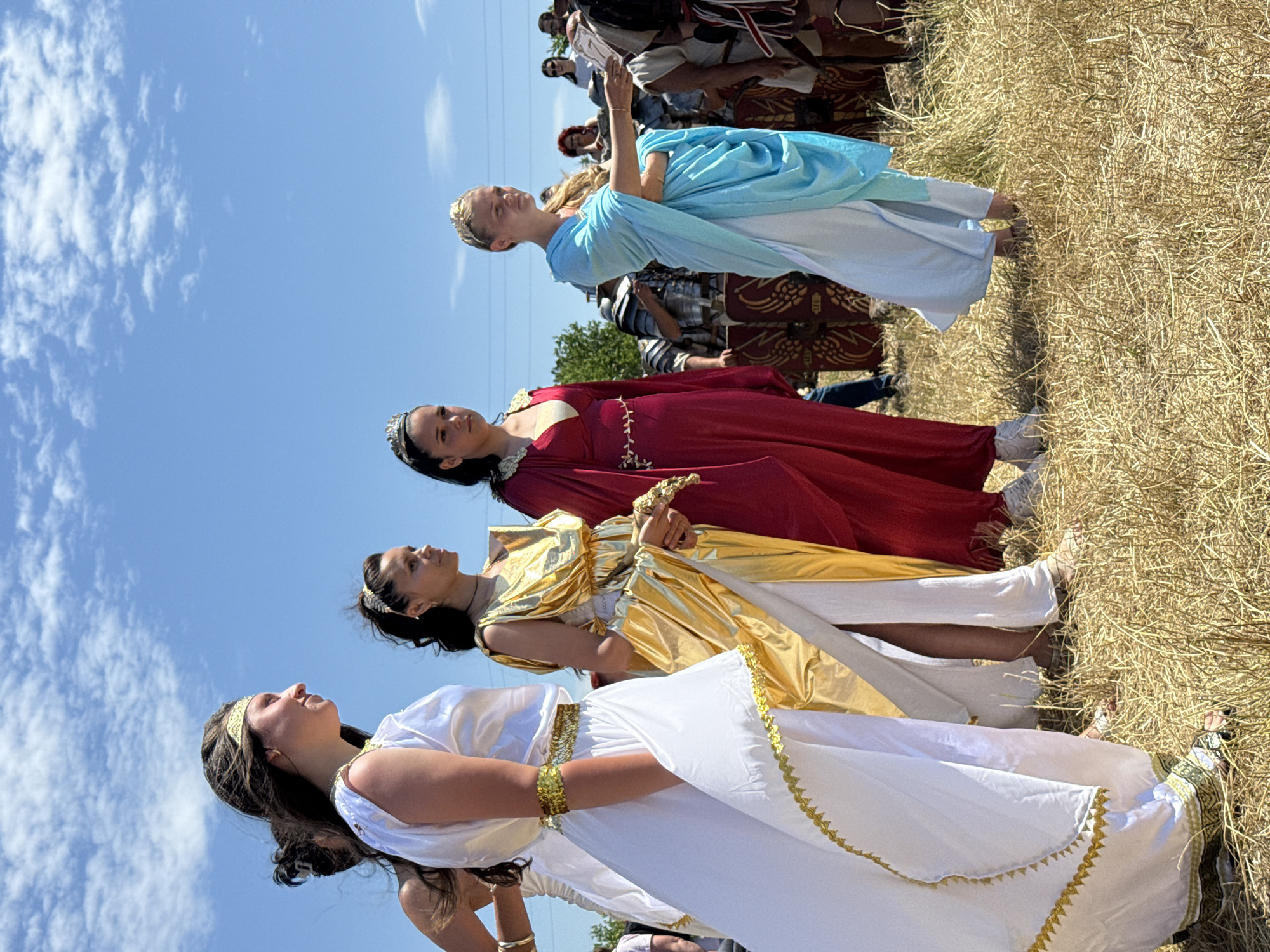 The image displays an outdoor scene in Gigen, Bulgaria, under a clear blue sky with scattered white clouds. Five young women, dressed in costumes reminiscent of ancient Roman or Greek attire, stand on dry, light brown grass, positioned from the left to the right side of the frame.
From left to right:
1. A woman's left bare shoulder, upper arm, and the back of her head with dark hair are partially visible, suggesting a light-colored garment.
2. A woman with long dark hair and a gold headband wears a floor-length white toga-style dress with gold trim along the neckline, armholes, and hem, and a gold upper arm band. She looks towards the right with a slight smile. Her feet are bare or in subtle sandals.
3. A woman with dark hair and a golden leaf-motif headband wears a flowing, floor-length gold-colored dress with a high slit revealing a white underskirt. She has a gold bracelet on her left wrist and holds a small, light-colored object in her hands, looking towards the right. Her feet are bare or in subtle sandals.
4. A woman with dark hair tied back, wearing a small silver tiara, is dressed in a long-sleeved, deep red flowing dress with a V-neckline. White sneakers are visible beneath her dress hem. She looks towards the right.
5. A woman with light hair, tied back, and a small gold tiara, wears a flowing light blue draped garment over a white dress. She extends her right hand, presenting a small white rectangular object. Her feet are bare or in subtle sandals.
In the mid-ground, several individuals are visible, including one person in modern clothing on the far left. Further right, multiple individuals in what appear to be Roman legionary armor, complete with metal breastplates and rectangular shields, are partially seen. The far background includes a crowd of spectators in modern attire, some under trees, with distinct individuals such as a woman with red hair wearing sunglasses and another woman in a white t-shirt. The ground is covered with sparse, dry, light brown grass, indicating a sunny, possibly warm, environment. The scene suggests a historical re-enactment or cultural festival.