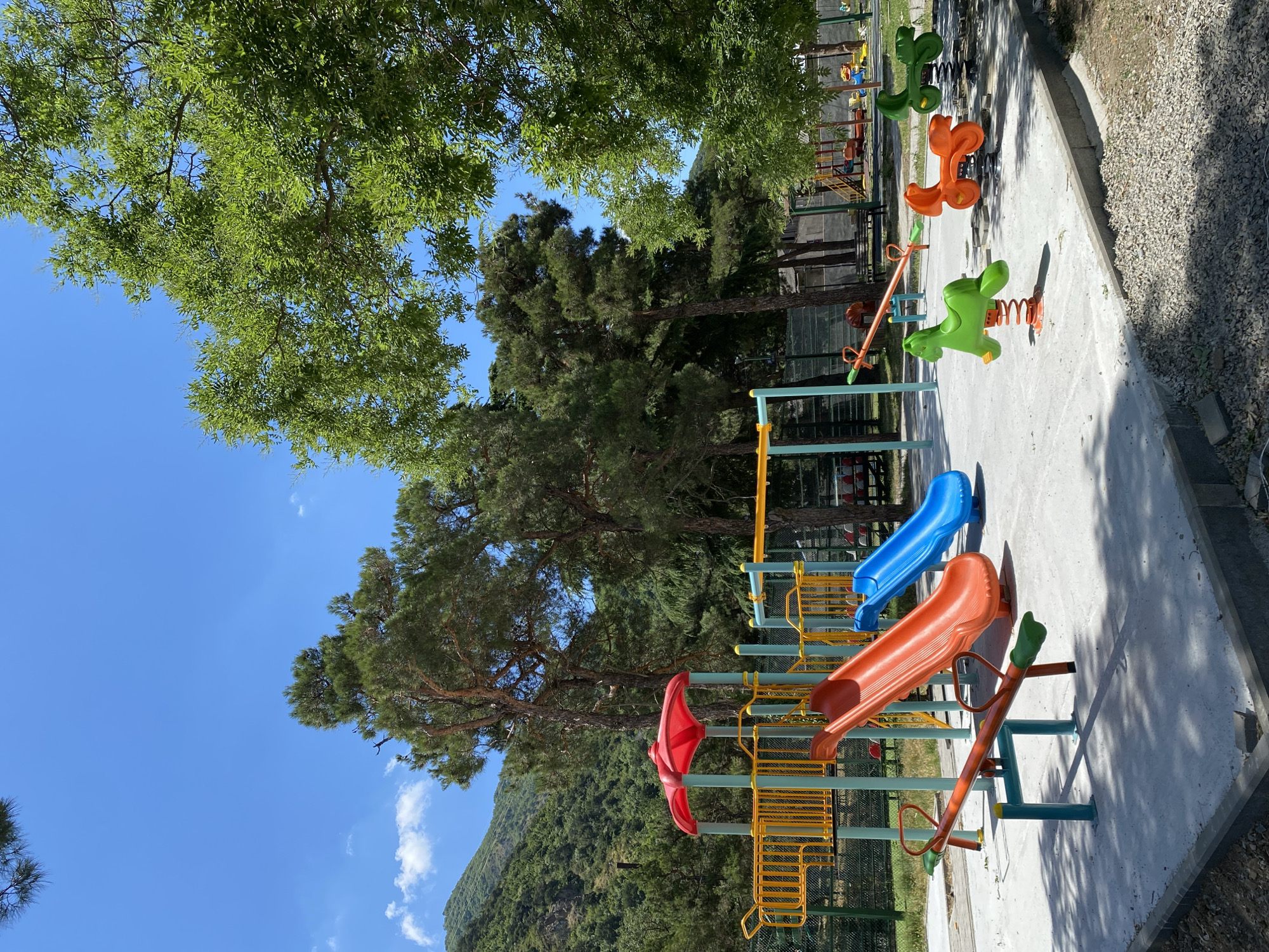 The image shows a playground in Mtskheta, Georgia, with a bright blue sky and green trees. The playground features several pieces of equipment, including orange and blue slides, a seesaw, and spring riders shaped like horses. In the background, a mountain covered in trees is visible. The ground is paved with concrete, and there are gravel areas around the playground. The scene is well-lit, suggesting it is daytime. There are no people visible in the image.