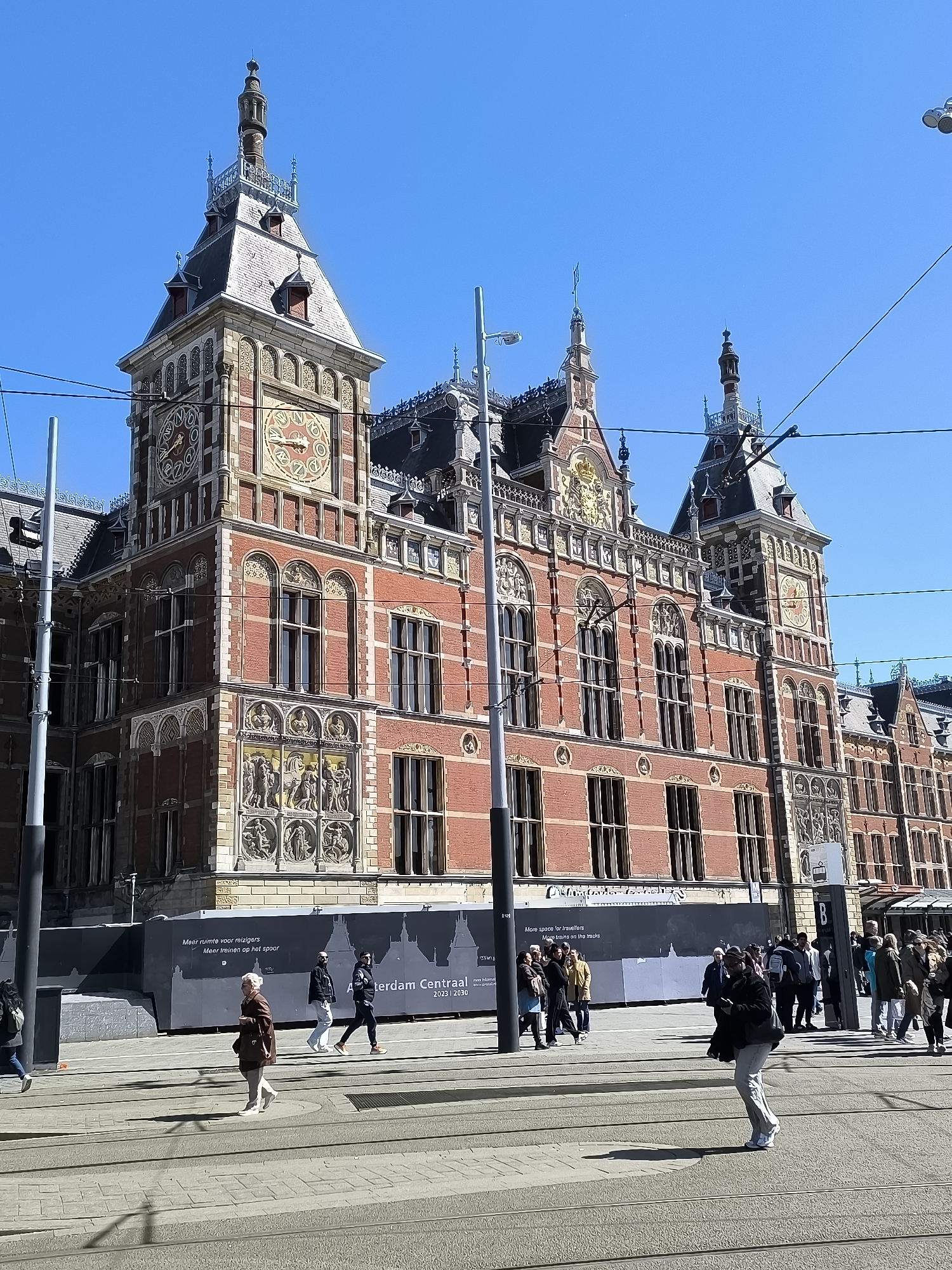 The image shows a street scene in Amsterdam, Netherlands, featuring the Amsterdam Centraal railway station. The station's facade, made of red brick, with ornate architectural details, is visible. The station features multiple clocks and sculptures. A construction barrier is partially obscuring the base of the building, displaying the text "Meer ruimte voor reizigers. Meer treinen op het spoor. Amsterdam Centraal. 2023 | 2030", with the translation "More space for travellers. More trains on the tracks". Several pedestrians are walking across the street in the foreground. The sky is clear and blue. There are overhead tram lines and lampposts visible. The ground is paved.