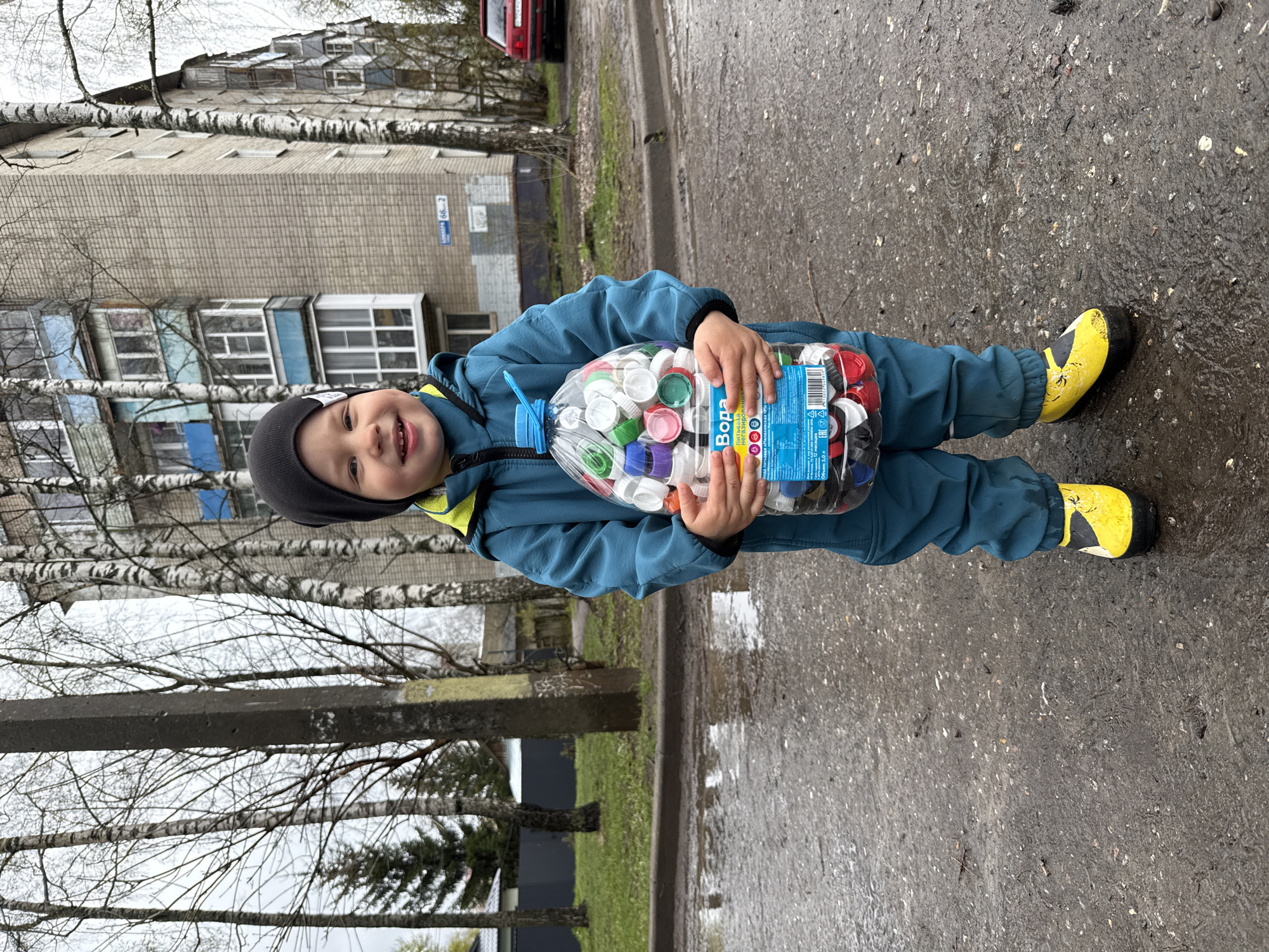 A young child, dressed in a blue jacket, matching overalls, a black beanie, and bright yellow boots, stands on a wet asphalt pavement. The child is smiling broadly at the camera, conveying a sense of happiness and pride. He is holding a large, transparent plastic bottle filled with a multitude of colorful plastic bottle caps. Visible text on the bottle includes "Вода" (Water), some smaller Russian text, and "Объем 5,0 л" (Volume 5.0 L).
The setting is an outdoor urban or suburban street. In the background, there is a large, multi-story apartment building, typical of Soviet-era architecture, with multiple windows and balconies. A blue street sign is affixed to the building, clearly displaying "ОКТЯБРЬСКАЯ 66 - 2" (Oktyabrskaya 66-2). Bare birch trees with white bark line the street, suggesting either late winter or early spring. The ground is visibly damp, possibly from recent rain, and reflects some light. A red car is partially visible further down the street on the left. The overall lighting suggests an overcast daytime sky. The child's activity appears to be proudly displaying his collection of bottle caps.